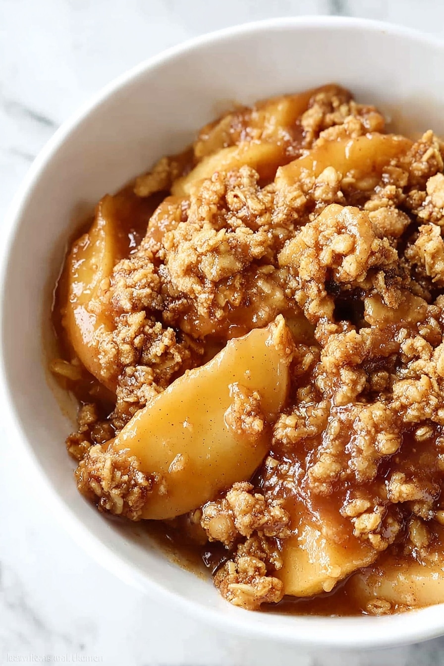A white bowl holds a warm apple dessert with three distinct layers: the bottom layer has soft, golden-brown apple slices coated in syrupy juices; above this is a crumbly, textured topping made of oats and brown sugar that looks crunchy and slightly toasted; the top layer is a smooth, creamy white scoop of melted vanilla ice cream that softly spreads over the crumble. A silver spoon rests in the bowl, partially visible at the back, all placed on a white marbled surface. Photo taken with an iphone --ar 2:3 --v 7 - Healthy Apple Crisp, healthy apple crisp recipe, wholesome apple dessert, gluten-free apple crisp, easy fall dessert