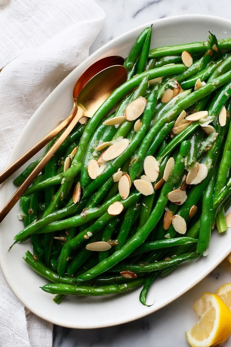 A white oval plate filled with bright green, shiny cooked green beans, arranged in a loose pile covering the plate. On top of the beans are thin, light tan almond slices scattered evenly, adding texture and contrast. Two small gold spoons rest on the beans, slightly overlapping, showing off the glossy surface of the veggies. The plate is placed on a white marbled surface, with a white cloth partially visible on the left side and a small piece of lemon on the right edge. The scene is well-lit, emphasizing the freshness and color of the dish. photo taken with an iphone --ar 2:3 --v 7 - Easy Green Beans Almondine, green beans almondine, green bean side dish, healthy green bean recipe, quick green bean recipes