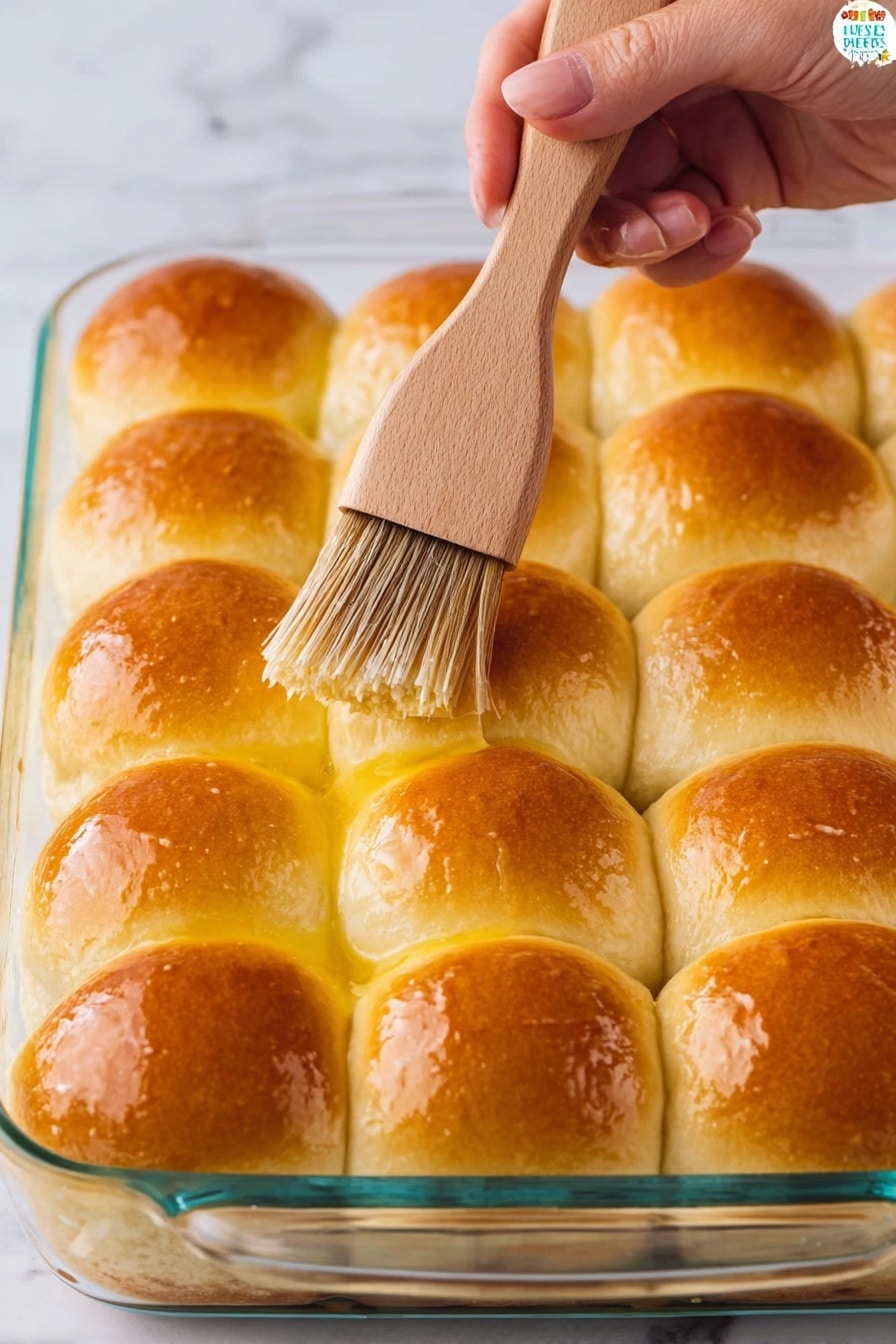 A glass baking dish holds a layer of 12 evenly arranged soft dinner rolls, each roll golden brown on top and shiny from being brushed with melted butter. The rolls sit closely together in a grid pattern with a soft, slightly fluffy texture. A woman's hand holds a light wooden brush with brown bristles, applying butter to the top of one roll. The background is a white marbled surface. photo taken with an iphone --ar 2:3 --v 7 - Fluffy Homemade Dinner Rolls, Soft dinner roll recipe, buttery dinner rolls, easy homemade bread, bakery-style dinner rolls