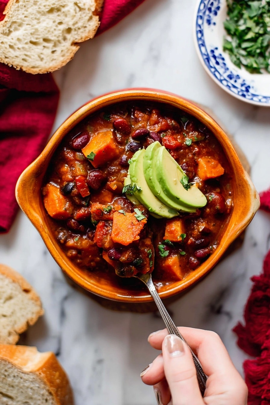A bowl of thick chili with visible layers of chunky tomato sauce, red and black beans, and soft, cubed orange sweet potatoes, topped with two slices of green avocado. The chili is served in a curved, orange bowl on a white marbled surface, with a woman's hand holding a spoon scooping into the chili. Surrounding the bowl are slices of bread on a white plate with blue patterns, a small bowl of chopped green herbs, and a red cloth in the background. Photo taken with an iphone --ar 2:3 --v 7 - Vegetarian Pumpkin Chili, hearty vegetarian chili, pumpkin chili recipe, vegetarian pumpkin soup, easy healthy chili