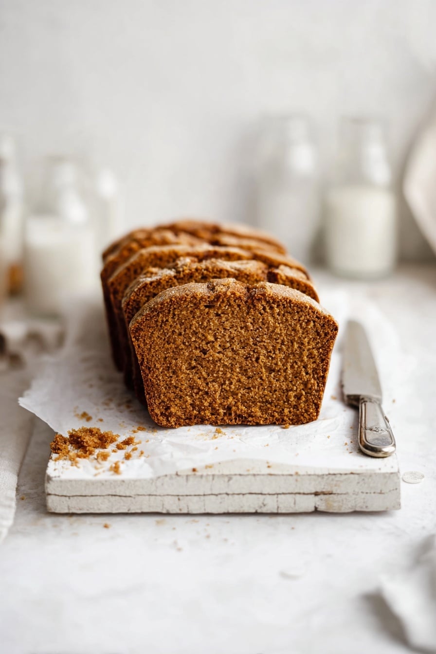 The image shows a loaf of brown bread sliced into six pieces, placed on white parchment paper on a rustic white wooden board. The bread has a golden-brown crust with a slightly rough texture and a dense, soft interior with small air holes. There are some crumbs scattered around the bread on the white marbled surface. To the upper right of the bread, there is a silver knife resting on top of a folded piece of white paper with text, and in the upper left, a clear empty glass and a white container are partially visible. Photo taken with an iphone --ar 2:3 --v 7 - Moist Pumpkin Bread, pumpkin bread recipe, easy pumpkin bread, autumn pumpkin bread, cozy pumpkin bread