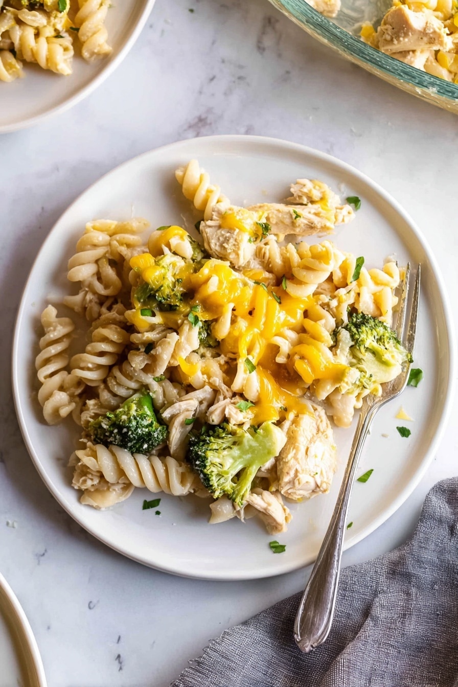 A white plate on a white marbled surface holds a serving of rotini pasta mixed with chunks of cooked chicken, bright green broccoli florets, and melted yellow cheese drizzled unevenly over the top. The pasta spirals are pale cream and slightly glossy, nestled closely with tender pieces of chicken and small green broccoli heads, all lightly sprinkled with small bits of fresh green herbs. A silver fork rests partially on the right edge of the plate, with its tines touching some pasta. There is a gray cloth napkin in the lower right corner and a partial view of another white plate and a glass baking dish in the background. Photo taken with an iphone --ar 2:3 --v 7 - Creamy Chicken Broccoli Noodle Casserole, cheesy chicken broccoli bake, easy comfort food dinner, hearty chicken noodle casserole, quick weeknight family meal