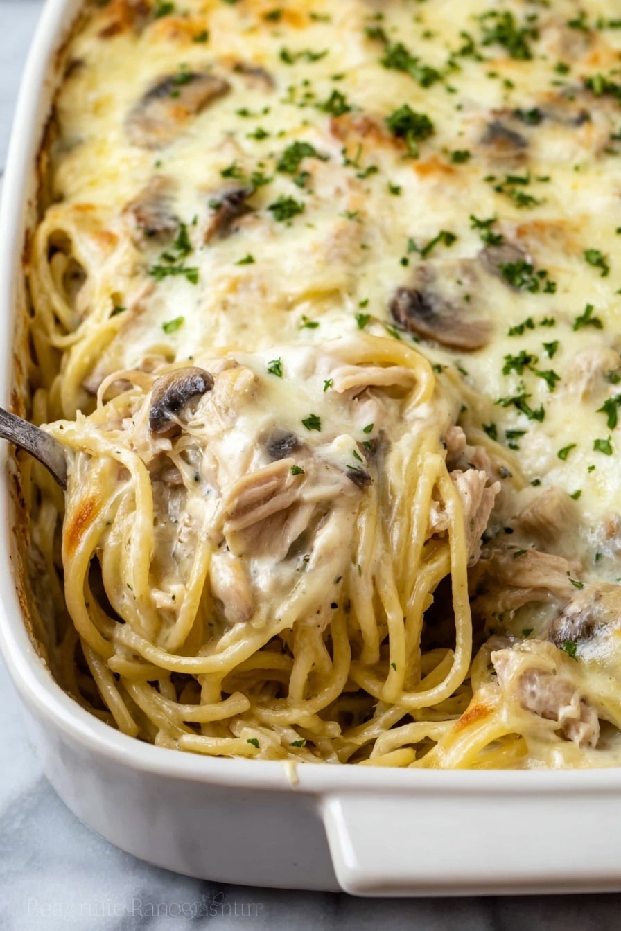 This dish shows a close-up view of a baked pasta casserole in a white ceramic dish. The casserole consists of creamy spaghetti mixed with pieces of light brown cooked meat and sliced mushrooms spread throughout. The top layer is covered with a smooth, melted white cheese sauce sprinkled with small bits of green herbs. A spoon is scooping some of the creamy pasta from the dish, showing the mixed layers of noodles, meat, and mushrooms inside. The dish is placed on a white marbled surface. photo taken with an iphone --ar 2:3 --v 7 - Creamy Chicken Tetrazzini Bake, chicken tetrazzini bake, creamy pasta casserole, easy chicken baked pasta, comfort food recipes