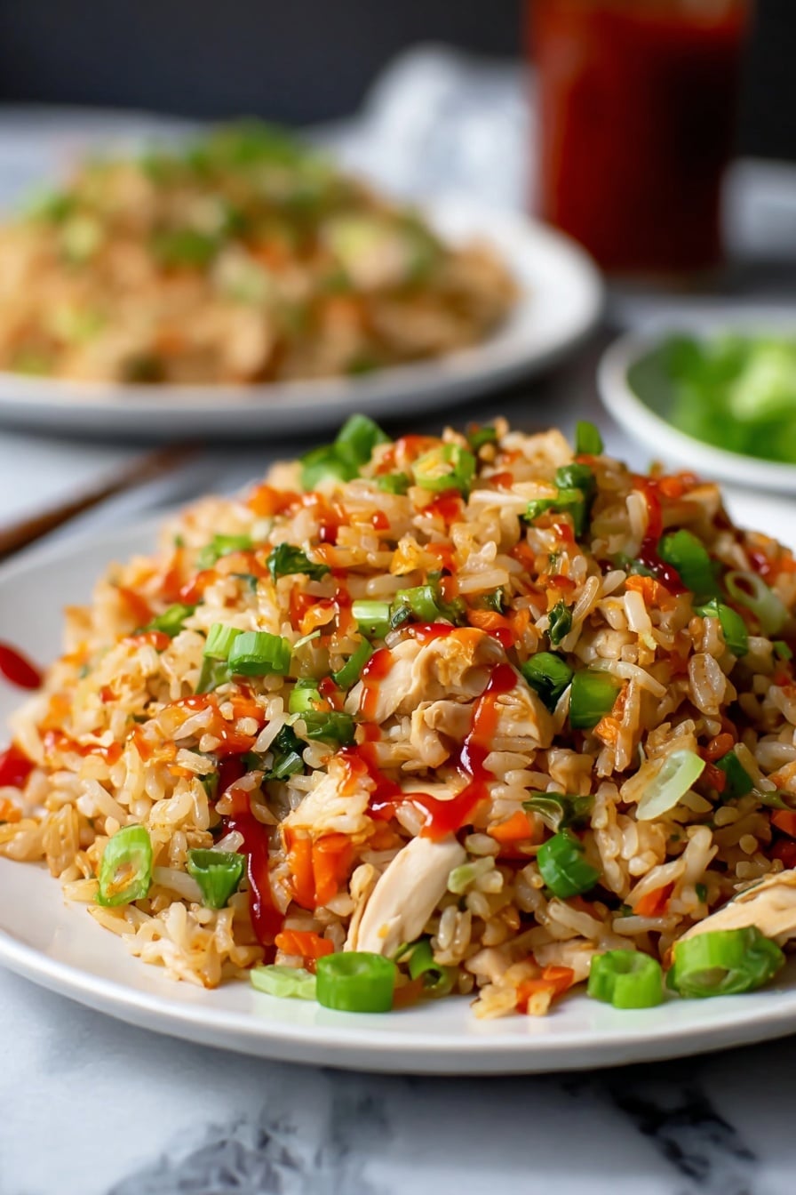 A close-up view of a white plate filled with fried rice, showing layers of light brown rice mixed with small pieces of white chicken, bright green chopped spring onions, and small orange-red peppers scattered throughout. The rice is lightly coated with red sauce drizzled on top, adding a glossy texture. In the background, there is a blurred white plate with more fried rice, placed on a white marbled surface. The overall colors are warm with green, red, and white contrasts. Photo taken with an iphone --ar 2:3 --v 7 - Easy Chicken Fried Rice, quick chicken fried rice, easy fried rice recipe, homemade chicken fried rice, best chicken fried rice