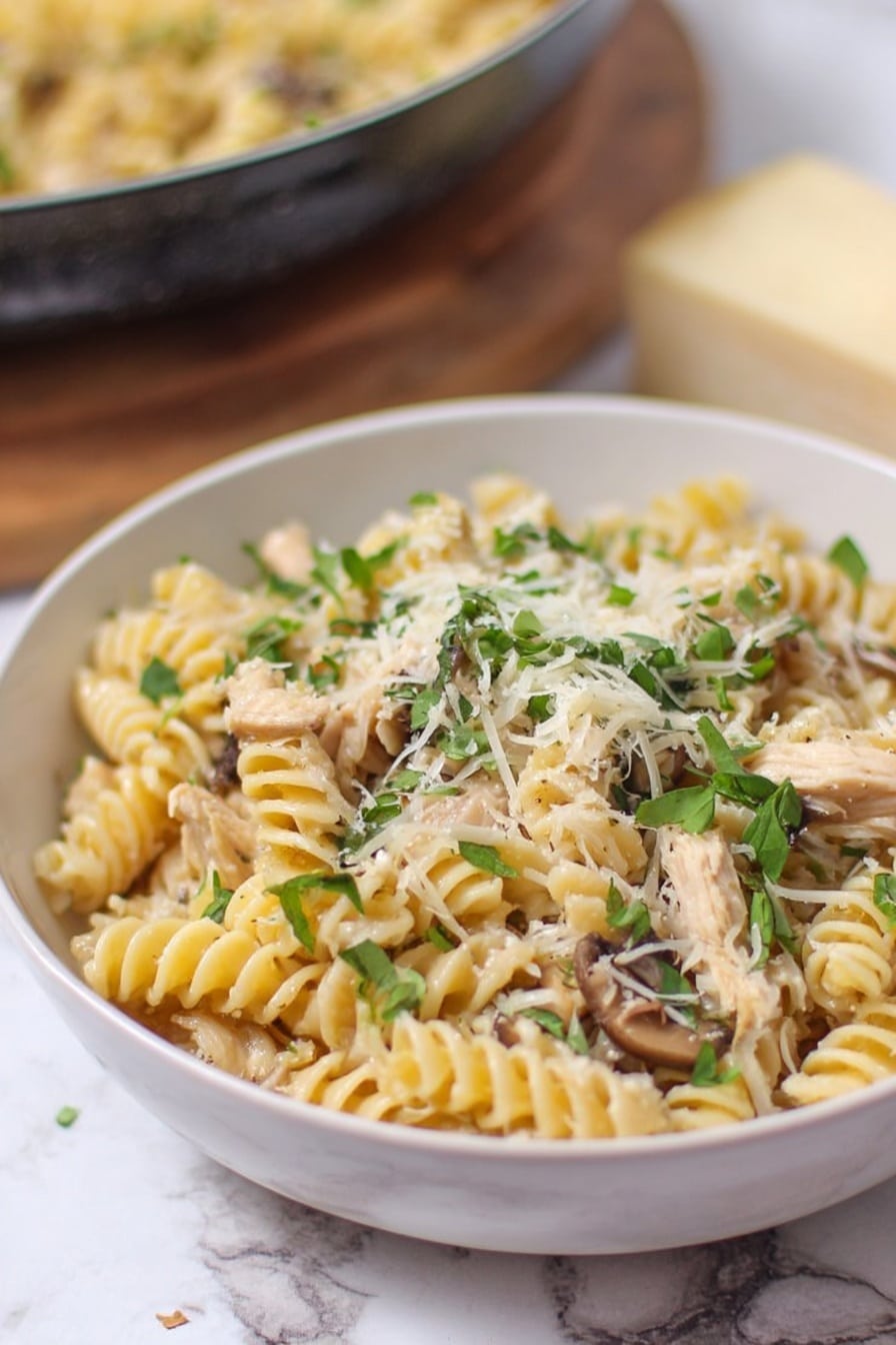 The image shows a white bowl filled with spiral pasta layered with light-colored shredded chicken and small bits of mushrooms. The top layer has finely grated cheese scattered evenly, which looks soft and slightly fluffy, and fresh green herb leaves spread all over, adding a fresh contrast. The background has a white marbled texture with a piece of cheese and a pan of more pasta slightly out of focus. The overall look is creamy and fresh, with natural light highlighting the details of the pasta and herbs. photo taken with an iphone --ar 2:3 --v 7 - Creamy Turkey Pasta, leftover turkey pasta, quick turkey pasta, easy creamy pasta, Thanksgiving turkey recipes
