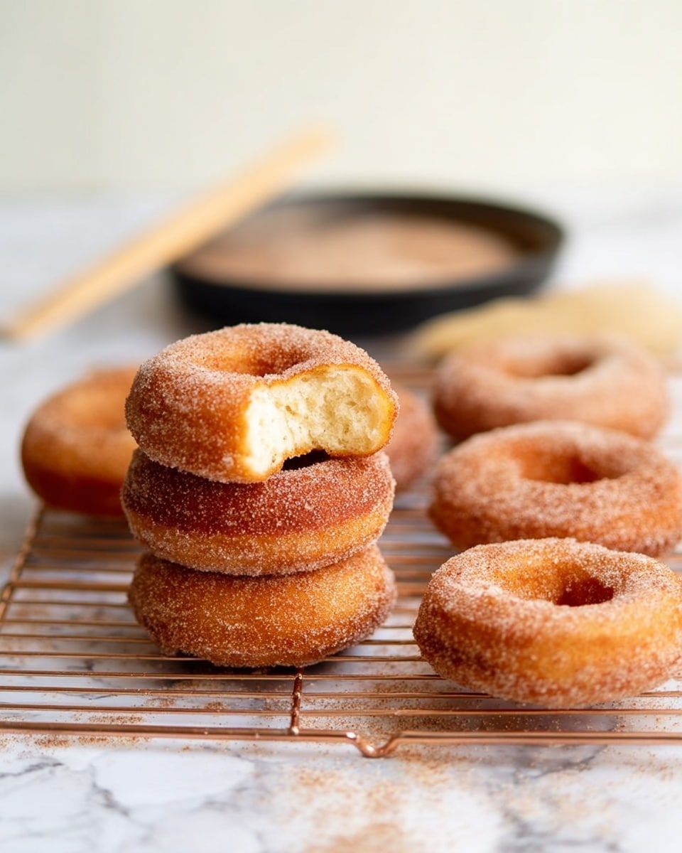 The image shows six cinnamon sugar-coated donuts placed on a metal cooling rack over a white marbled surface. Five whole round donuts sit flat on the rack, each with a rough, textured surface covered evenly in cinnamon sugar, giving them a warm brown color. One donut is held upright, showing a soft, pale beige inside with a visible bite taken out, revealing its fluffy texture. To the top right, there is a round metal pan filled with cinnamon sugar and a wooden pastry brush resting on its edge, its bristles dusted with sugar. The overall scene has a warm, inviting feel with a focus on the donuts' texture and sugar coating, photo taken with an iphone --ar 4:5 --v 7