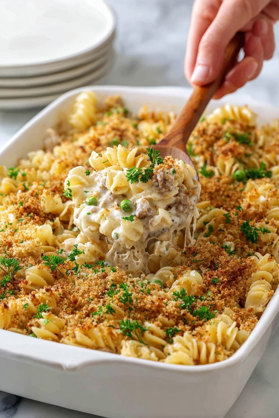 A white baking dish filled with a creamy pasta casserole made of curly pasta mixed with a white sauce, small pieces of light-colored meat, and green peas. The top layer is covered with a golden-brown crunchy breadcrumb topping, sprinkled with fresh green parsley pieces. At the front, a wooden spoon with a woman's hand holding it scoops out a portion, showing the creamy inside with visible pasta, meat, and peas. The dish sits on a white marbled surface with a stack of white plates blurred in the background. photo taken with an iphone --ar 2:3 --v 7 - Classic Tuna Noodle Casserole, Tuna Noodle Casserole with Parmesan Breadcrumb Topping, Easy Tuna Casserole Recipe, Creamy Tuna Casserole, Comfort Food Tuna Bake