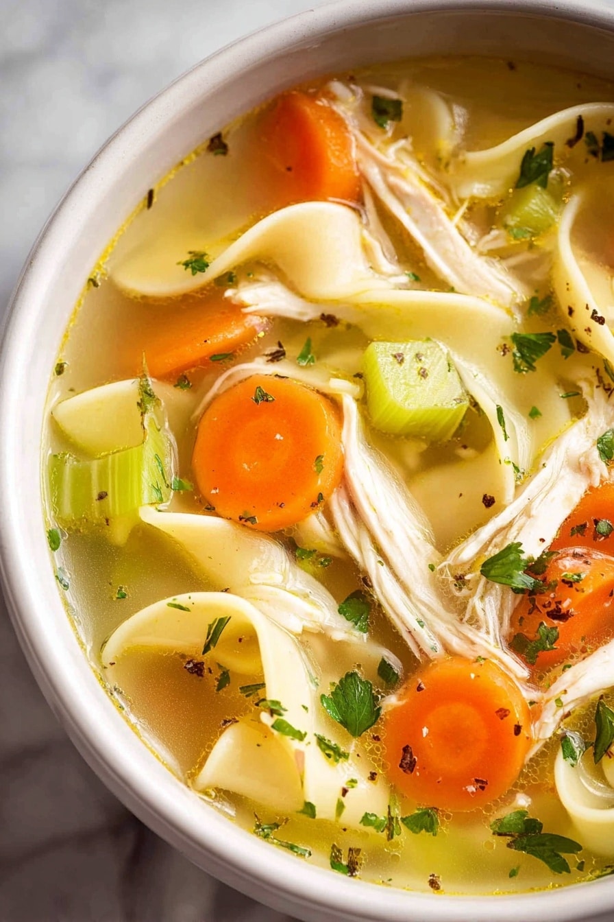 A white bowl filled with clear golden broth containing long, flat yellow noodles, small orange carrot cubes, and green celery pieces. Shredded light beige chicken is spread evenly throughout, with flecks of green herbs and black pepper for extra color. A single dark brown bay leaf is visible near the bottom, and a silver spoon is partially submerged in the soup. The bowl rests on a white marbled surface with a small white cup of chopped green herbs and another white cup with a white creamy substance in the background, photo taken with an iphone --ar 2:3 --v 7 - Homemade Chicken Noodle Soup from Scratch, chicken noodle soup, homemade chicken soup, comforting chicken noodle recipe, fresh chicken soup