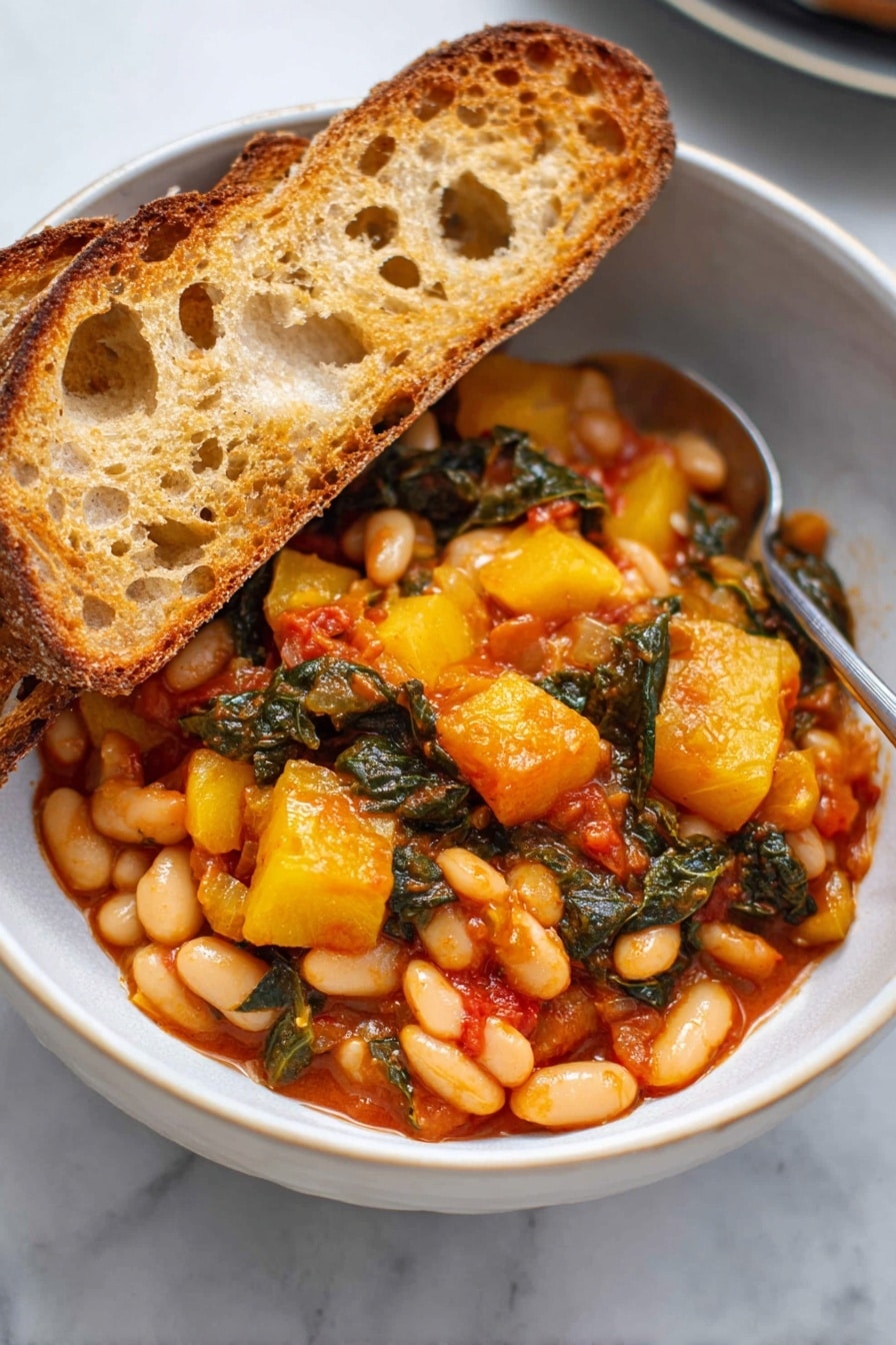 A white bowl filled with two thick slices of toasted bread resting on the left side, showing their golden-brown crispy texture with some holes and rough edges. The main part of the bowl is filled with a colorful mix of cooked white beans, yellow chunks of squash, and dark green leafy vegetables, all coated in a rich, slightly shiny red-orange sauce. The ingredients are close together, showing a hearty, rustic stew. A silver spoon is partially visible on the right side of the bowl, touching the food. The bowl sits on a white marbled surface. photo taken with an iphone --ar 2:3 --v 7 - Stewed Great Northern Beans with Kale Tomatoes and Herb-Infused Broth, hearty bean and kale stew, healthy vegetarian bean recipe, nourishing winter dinner idea, easy bean and greens recipe