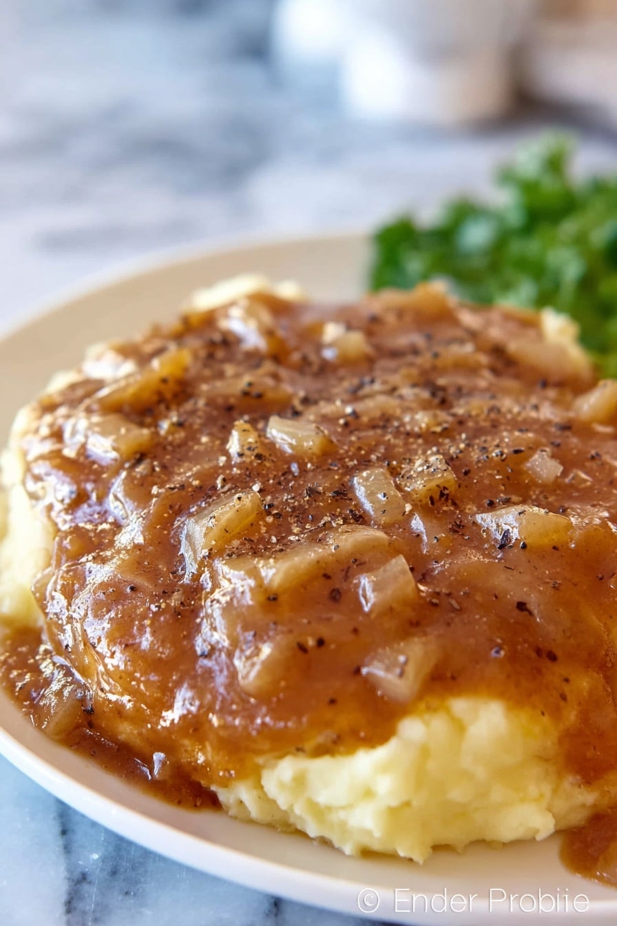 A white plate with mashed potatoes as the base layer, creamy and light yellow in color with a smooth texture, topped with a thick brown gravy that has small diced onion pieces spread evenly across the surface. The gravy covers a large piece of meat underneath, sprinkled with black pepper flakes. The background shows an out-of-focus white marbled surface and some green garnish in the back. Photo taken with an iphone --ar 2:3 --v 7 - Crock Pot Cube Steak, slow cooker cube steak recipe, easy cube steak dinner, tender beef stew, hearty slow cooker meals