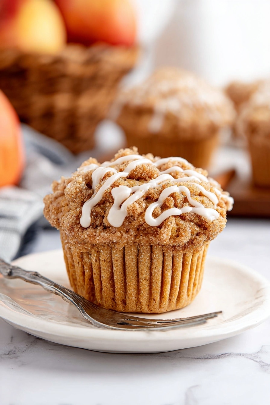 A single muffin sits centered on a white plate with a silver fork resting next to it. The muffin has a light brown, soft textured base with a crumbly streusel topping in a darker brown shade. White icing drizzle runs in thin lines over the streusel, adding contrast. The plate and muffin are placed on a white marbled surface. In the background, blurred muffins and a basket of apples add warm tones but stay out of focus. Photo taken with an iphone --ar 2:3 --v 7 - Cinnamon Apple Muffins with Crumble Topping and Glaze, apple muffin recipes, fall breakfast muffins, easy apple muffins, cozy baking recipes