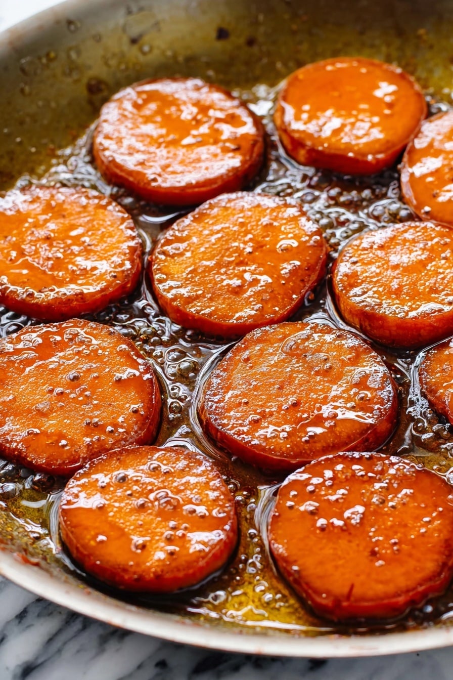 The image shows a white oval dish filled with thick, round slices of cooked sweet potatoes. The sweet potato slices are covered in a shiny, dark caramel-like sauce that makes them look glossy and soft. The slices are arranged closely together, slightly overlapping, and their orange color is rich and warm, enhanced by the dark syrup. The sauce pools around the edges of the slices, filling the bottom of the dish, and the texture looks smooth and sticky. The setting is on a white marbled surface. photo taken with an iphone --ar 2:3 --v 7 - Bourbon Glazed Candied Sweet Potatoes, sweet potato side dish with bourbon, holiday sweet potato recipes, easy candied sweet potatoes, flavorful sweet potato dish