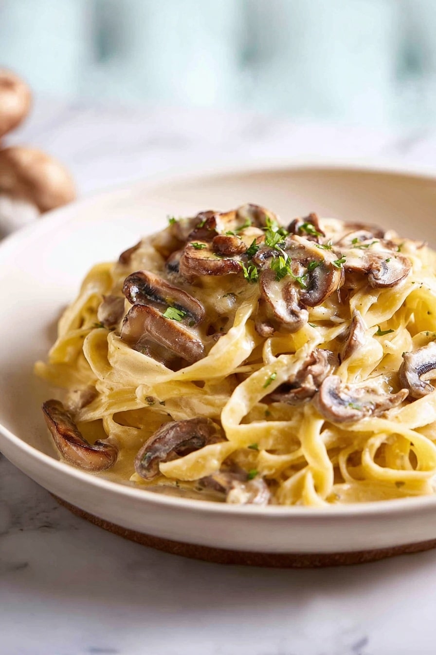 The image shows a mound of creamy fettuccine pasta on a plain white plate. The pasta is light yellow and looks soft and smooth, with thin sauce coating each strand. On top and mixed through are brown sliced mushrooms with a slight gloss, adding texture and earthy colors. Small green herb bits are sprinkled over the pasta, giving little pops of color. The plate sits on a white marbled surface, with a blurred background that is softly lit showing a light blue and white shade. Photo taken with an iphone --ar 2:3 --v 7 - Creamy Mushroom Pasta, Mushroom Pasta Recipe, Easy Mushroom Pasta, Vegetarian Mushroom Pasta, Comfort Food Pasta