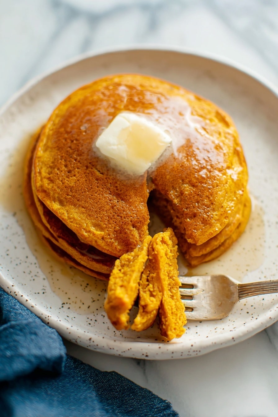 There is a white plate with three golden-brown pancakes stacked slightly unevenly with a clear textured top pancake showing tiny holes. A gold fork rests on the top edge of the plate. Next to the plate on the white marbled surface, there is a pile of more pancakes on another white plate that shows their soft, porous texture. A small white jug with dark syrup is near the plates, and a blue cloth is placed on the surface beside the plate. The photo taken with an iphone --ar 2:3 --v 7 - Whole Wheat Pumpkin Pancakes, pumpkin breakfast ideas, healthy autumn pancakes, cozy fall brunch recipes, fluffy pumpkin pancakes