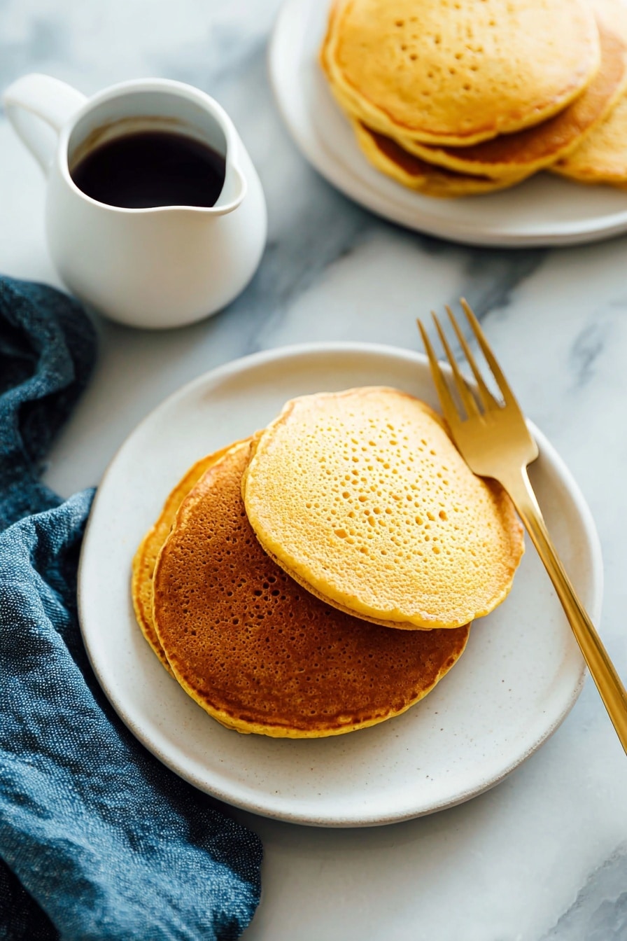 There is a stack of three golden-brown pancakes on a white plate. The pancakes have a slightly porous texture and are arranged with the largest at the bottom and the smallest on top. A light amber syrup is poured over the top pancake, dripping down the sides and pooling a little on the plate. A silver fork rests under the stack, on the left side, and the plate sits on a white marbled surface with a blue cloth nearby. The lighting is soft and natural, highlighting the warm tones of the pancakes and syrup. photo taken with an iphone --ar 2:3 --v 7 - Whole Wheat Pumpkin Pancakes, pumpkin breakfast ideas, healthy autumn pancakes, cozy fall brunch recipes, fluffy pumpkin pancakes