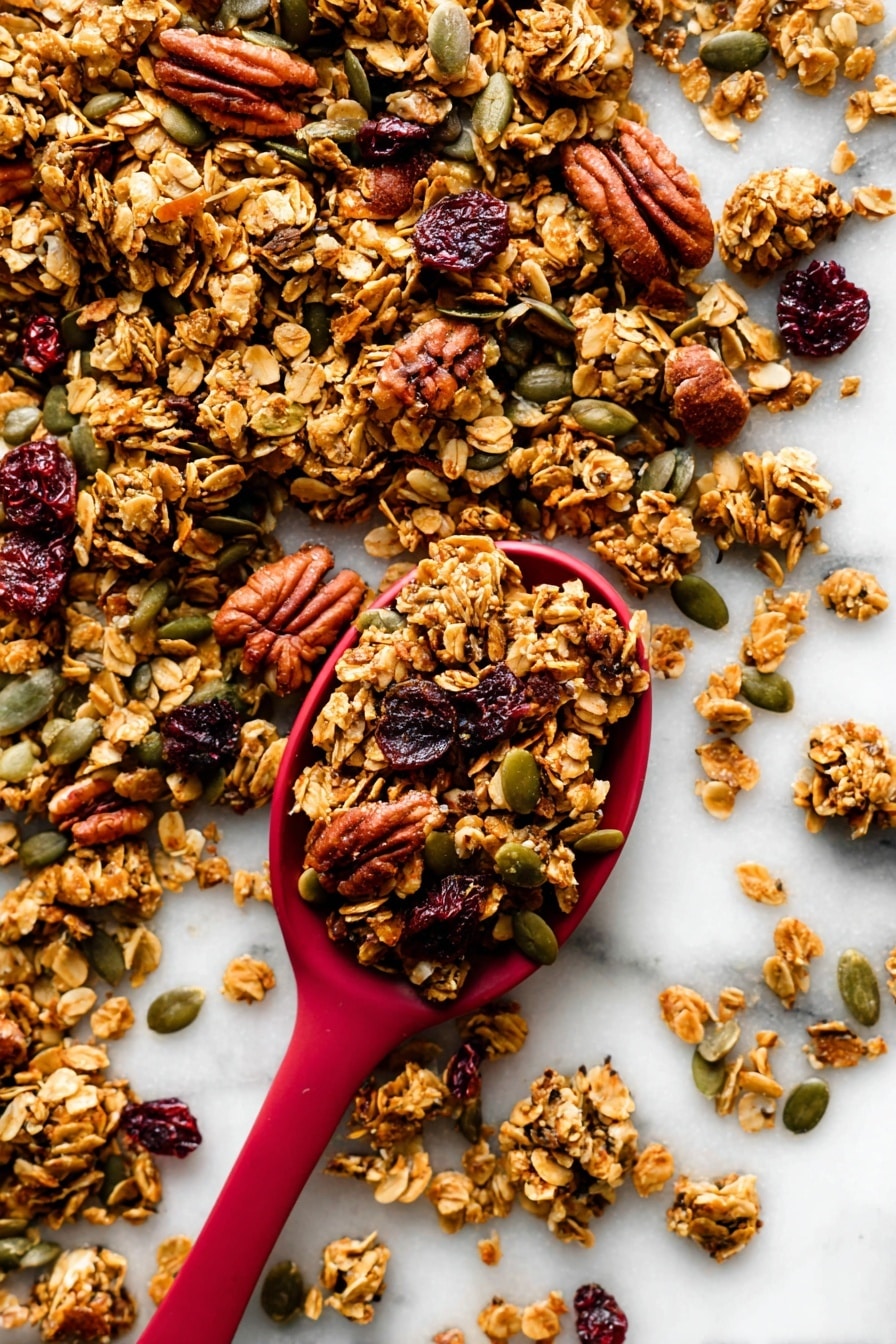 The image shows a close-up of crunchy granola scattered on a white marbled surface with a red spoon filled with granola placed near the center. The granola is made of golden-brown toasted oat clusters mixed with darker brown pecans, green pumpkin seeds, and dark red dried cranberries. The textures vary from rough and chunky oats and nuts to smooth seeds, creating a rich and inviting look. The red spoon contrasts strongly with the white background and the natural colors of the granola around it. photo taken with an iphone --ar 2:3 --v 7 - Pumpkin Spice Granola with Cranberries, fall granola recipe, pumpkin spice breakfast, healthy pumpkin granola, crunchy cranberry granola