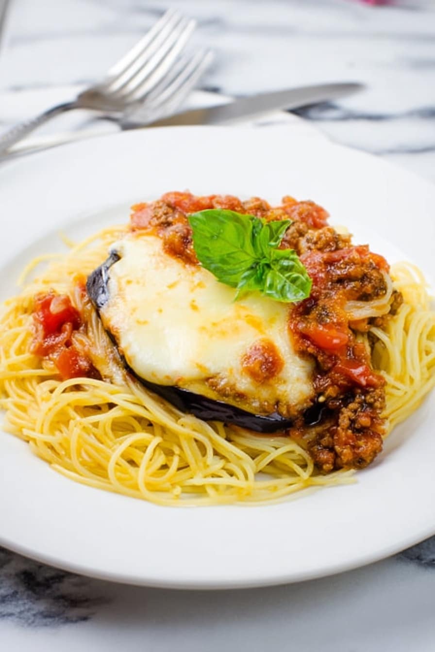 A white plate holds a dish with a bottom layer of thin yellow spaghetti noodles spread evenly. On top of the noodles are two thick slices of eggplant covered with a melted, light golden layer of cheese that looks soft and slightly browned in spots. A chunky meat and tomato sauce with visible small pieces of tomato and meat is spooned over the eggplant slices. A fresh green basil leaf is placed on top as garnish. The plate sits on a white marbled surface with a fork and knife just visible at the bottom left corner. Photo taken with an iphone --ar 2:3 --v 7 - Eggplant Parmesan Spaghetti with Meat Sauce, Italian eggplant spaghetti, baked eggplant pasta, hearty meat sauce recipes, crispy eggplant pasta dish
