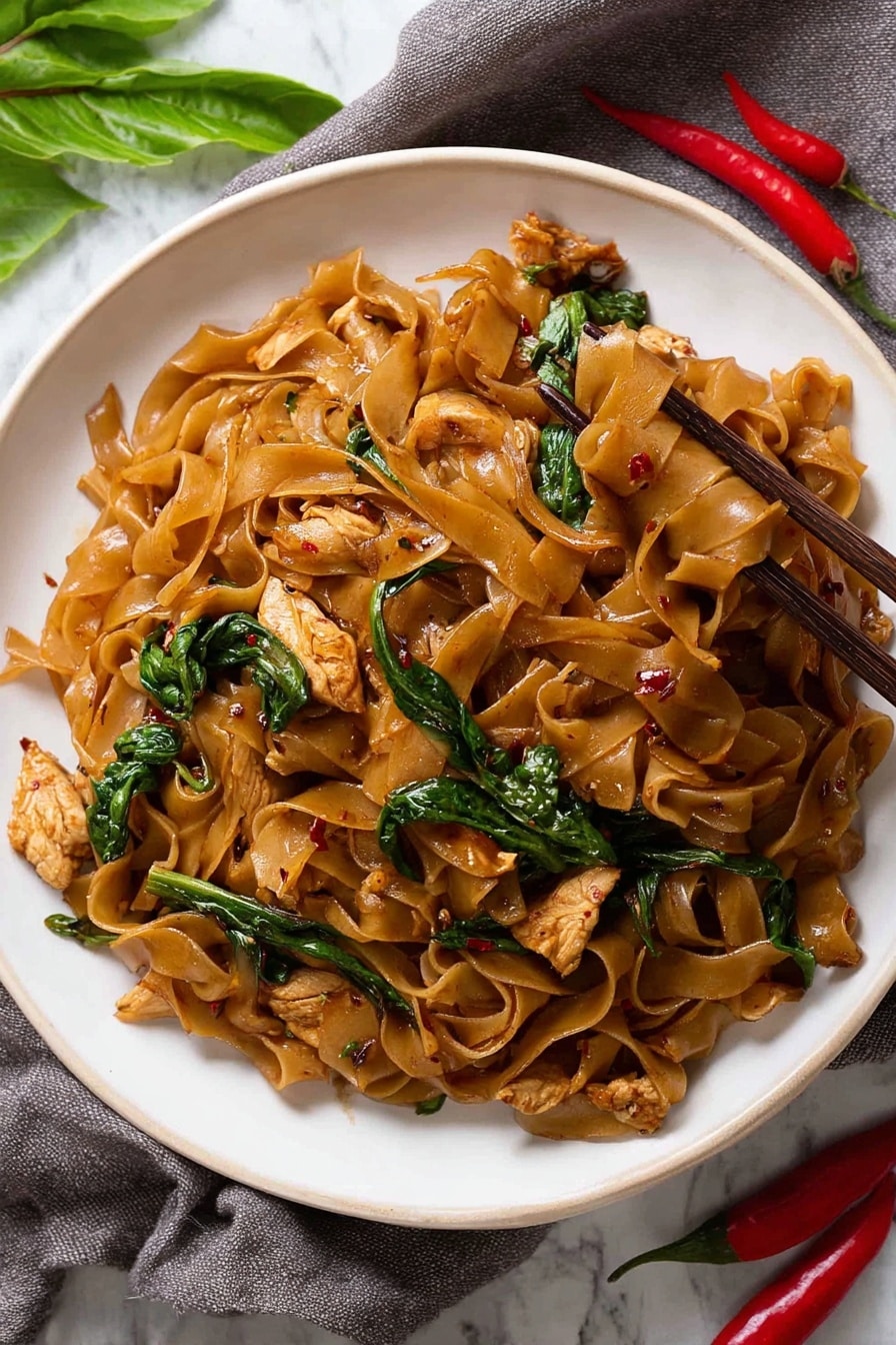 The dish shows a white round plate filled with wide, flat brown noodles mixed with pieces of cooked light brown chicken and green leafy vegetables scattered throughout. The noodles have a shiny, slightly oily texture, with small red chili flakes visible on some parts, giving a spicy look. The green vegetables add a fresh pop of color contrasting the darker noodles. Brown chopsticks are placed on the right edge of the plate, slightly lifting some noodles. The plate sits on a white marbled surface with a gray cloth and red chili peppers and green leaves partially visible in the background. Photo taken with an iphone --ar 2:3 --v 7 - Spicy Thai Drunken Noodles, Thai street food noodles, spicy Thai noodles recipe, authentic Thai drunken noodles, quick Thai noodle dish