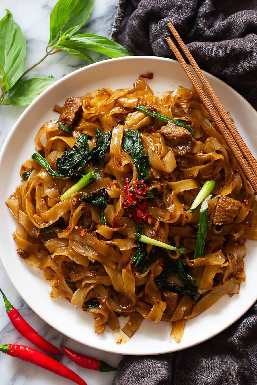 A white plate filled with flat brown noodles mixed with glossy pieces of cooked meat that have a caramel color. Scattered through the noodles are bright green pieces of vegetables, including spring onions with white and green parts, and dark leafy greens, all coated in a slightly oily sauce with hints of red chili flakes. A pair of wooden chopsticks rests on the edge of the plate, gripping some noodles and meat. The plate sits on a white marbled surface with a dark grey cloth draped underneath and small bright red chili peppers with fresh green leaves nearby. Photo taken with an iphone --ar 2:3 --v 7 - Spicy Thai Drunken Noodles, Thai street food noodles, spicy Thai noodles recipe, authentic Thai drunken noodles, quick Thai noodle dish