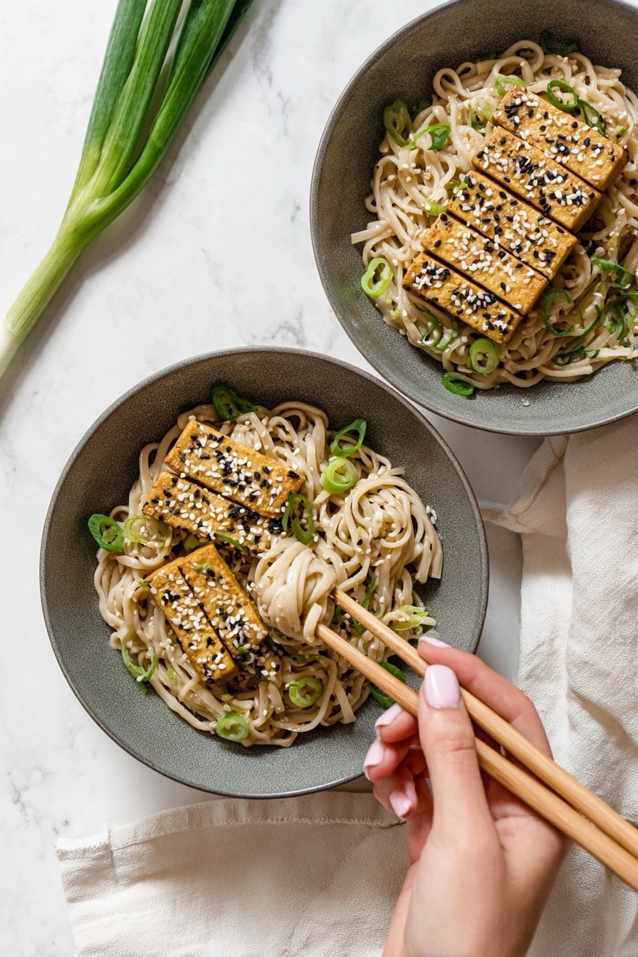 A round white bowl filled with creamy light beige noodles mixed with small green pieces, topped with four rectangular golden brown tofu slices sprinkled with white and black sesame seeds and chopped green onions. The noodles have a smooth texture, and the tofu pieces are placed side by side in the center. The bowl is set on a white marbled surface with two whole green onions placed above it and a pair of wooden chopsticks resting on a beige cloth to the right. Photo taken with an iphone --ar 2:3 --v 7 - Peanut Udon Noodles, Thai peanut udon, quick udon noodle recipe, vegan peanut noodles, easy Asian noodle dish