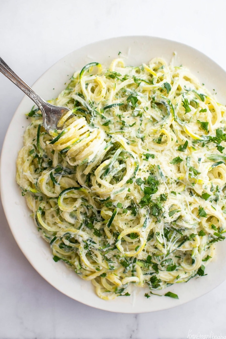 A white plate filled with a creamy dish made of spiral-shaped green and light yellow vegetable noodles, mixed with a white creamy sauce that coats the noodles evenly. There are small green herb leaves sprinkled throughout the dish, adding a fresh look. A silver fork is twirling some of the noodles in the top left area of the plate. The plate rests on a white marbled surface. photo taken with an iphone --ar 2:3 --v 7 - Zucchini Alfredo, healthy zucchini Alfredo, low carb Alfredo with zucchini, creamy zucchini pasta, quick Zucchini Alfredo dinner