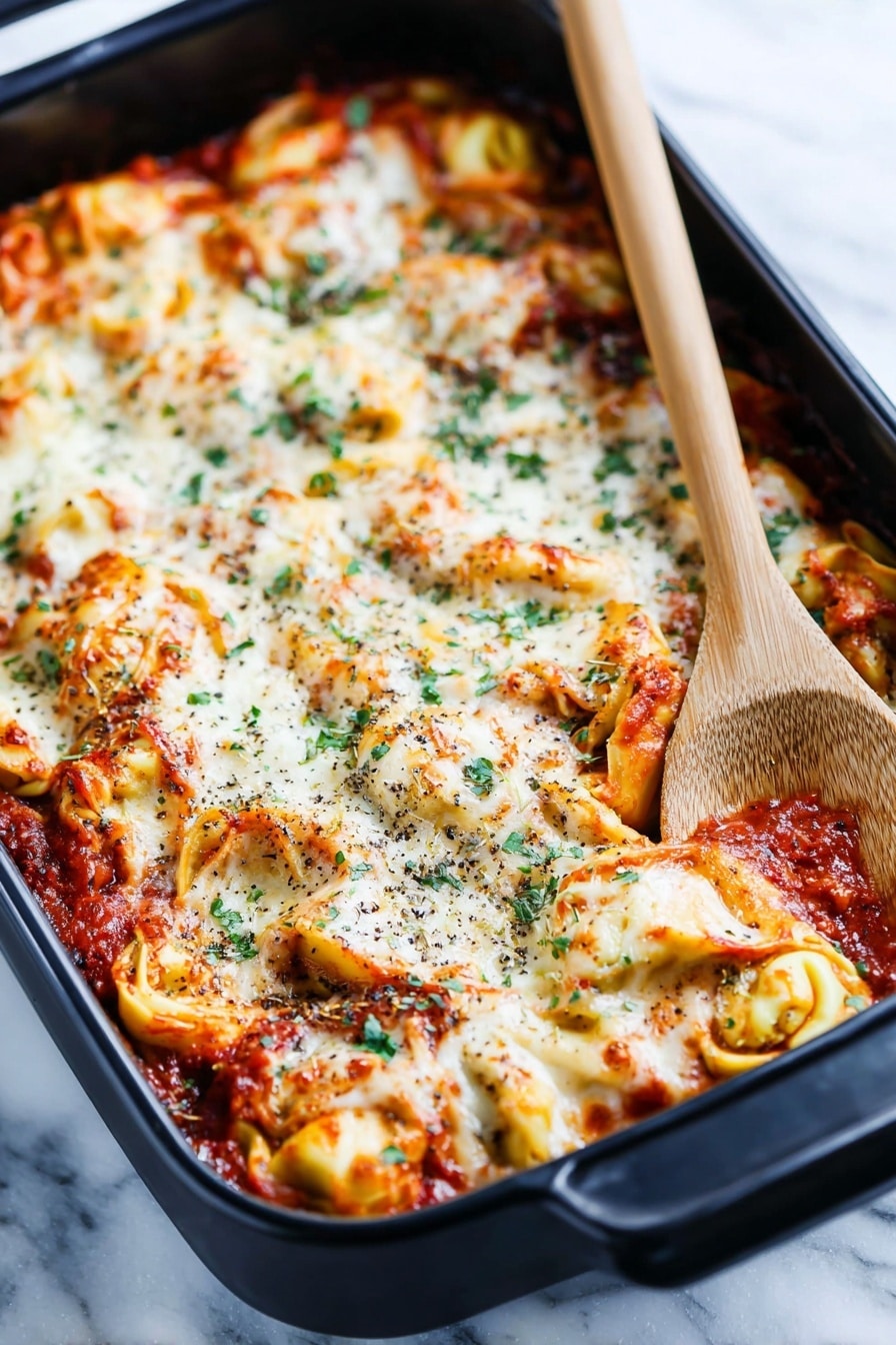 A black baking dish holds a layered tortellini pasta bake with a rich tomato sauce. The top layer is melted white cheese, bubbly and slightly browned in spots, mixed with twisted yellow tortellini filled pasta visible underneath. The sauce is deep red with bits of herbs and small chunks of meat mixed in. Scattered green herb leaves add color contrast across the top. The dish rests on a white marbled surface with a folded striped cloth nearby and a wooden-handled black spoon on the right edge of the frame, creating a cozy kitchen feel. photo taken with an iphone --ar 2:3 --v 7 - Cheesy Baked Tortellini Casserole, cheesy tortellini casserole, baked tortellini recipe, easy tortellini bake, comforting pasta casserole
