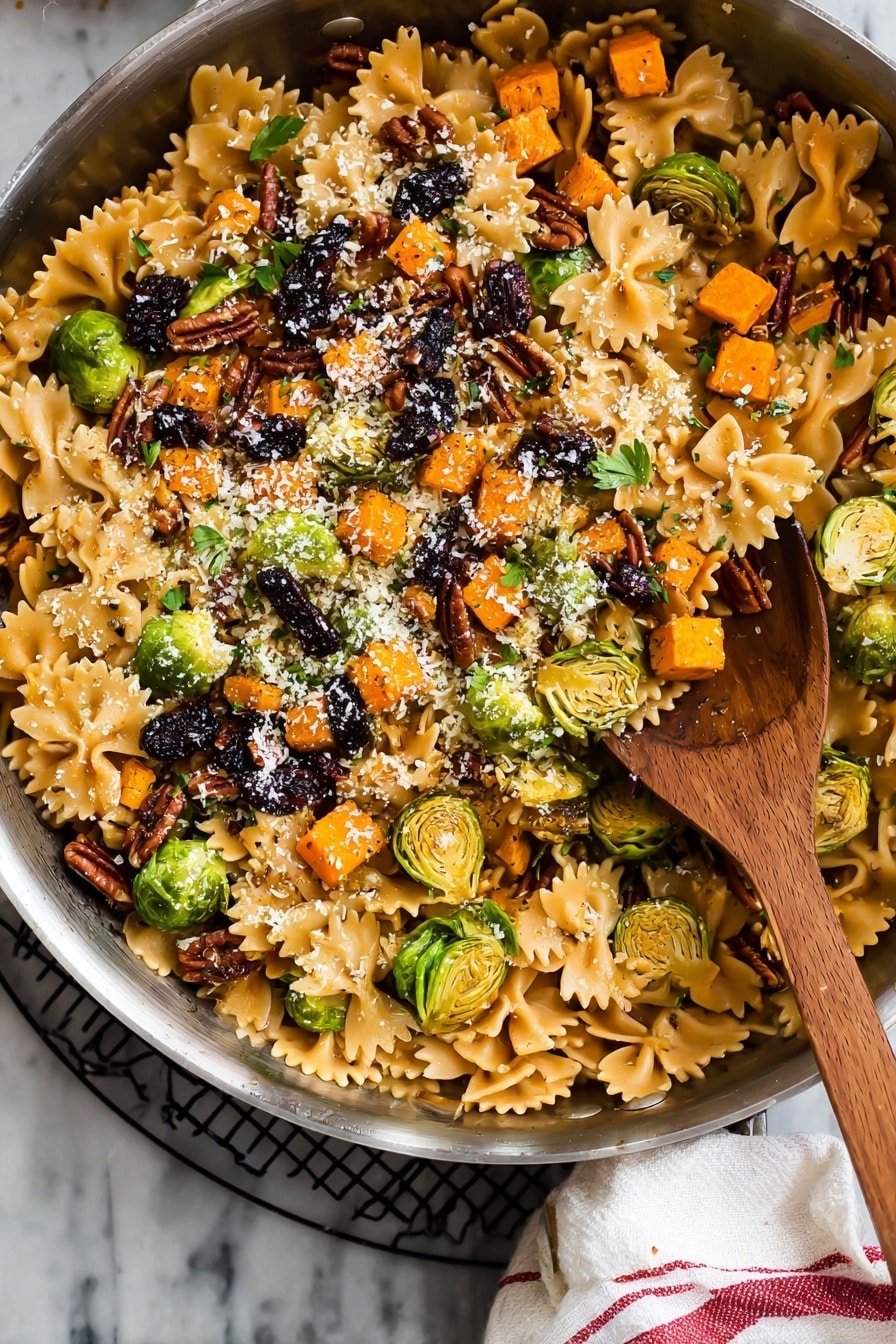 A large silver pan filled with a colorful pasta dish featuring three layers: the bottom layer has light brown farfalle pasta with smooth, slightly shiny texture; the middle layer contains bright orange small cubed roasted vegetables and sliced green brussels sprouts, some with charred marks, scattered evenly; the top layer is sprinkled with chopped dark brown pecans, black dried fruit pieces, small green herb leaves, and a light dusting of finely grated white cheese. A wooden spoon with a rounded bowl sits inside the pan. The pan rests on a black wire rack with a white cloth with red stripes on the side, all set on a white marbled surface. Photo taken with an iphone --ar 2:3 --v 7 - Brussels Sprouts Pasta with Brown Butter, Brussels sprouts pasta, fall pasta recipes, cozy vegetable pasta, caramelized Brussels sprouts recipes
