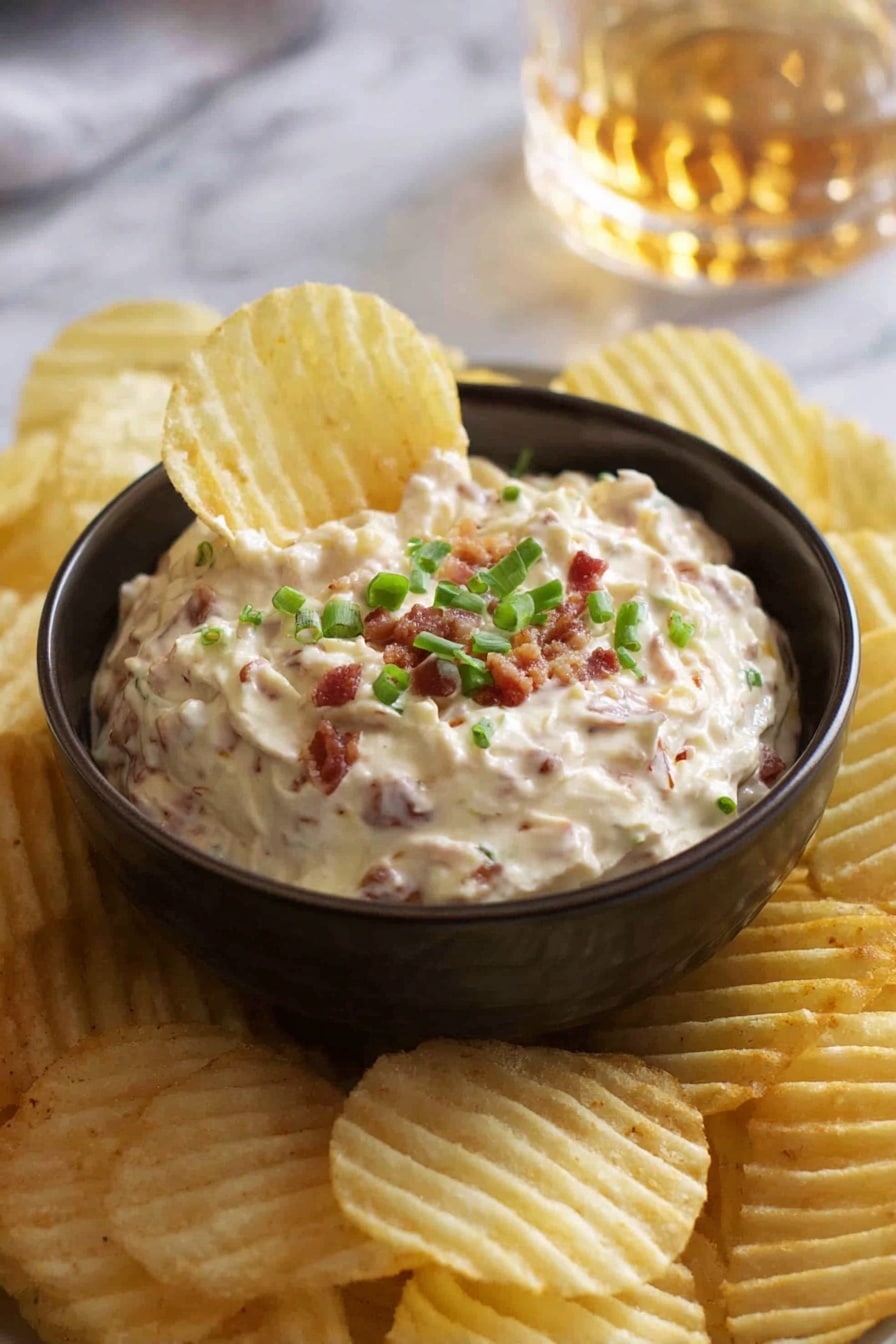 A small dark bowl filled with creamy dip that has small pieces of bacon mixed in, topped with chopped green onions. The bowl sits on a white marbled surface and is surrounded by many ridged potato chips, one chip is dipped into the bowl showing the texture of the dip. In the background, there is a blurred glass with a golden liquid. Photo taken with an iphone --ar 2:3 --v 7 - Homemade French Onion Dip, French Onion Dip recipe, caramelized onion dip, creamy onion dip, party dip recipes
