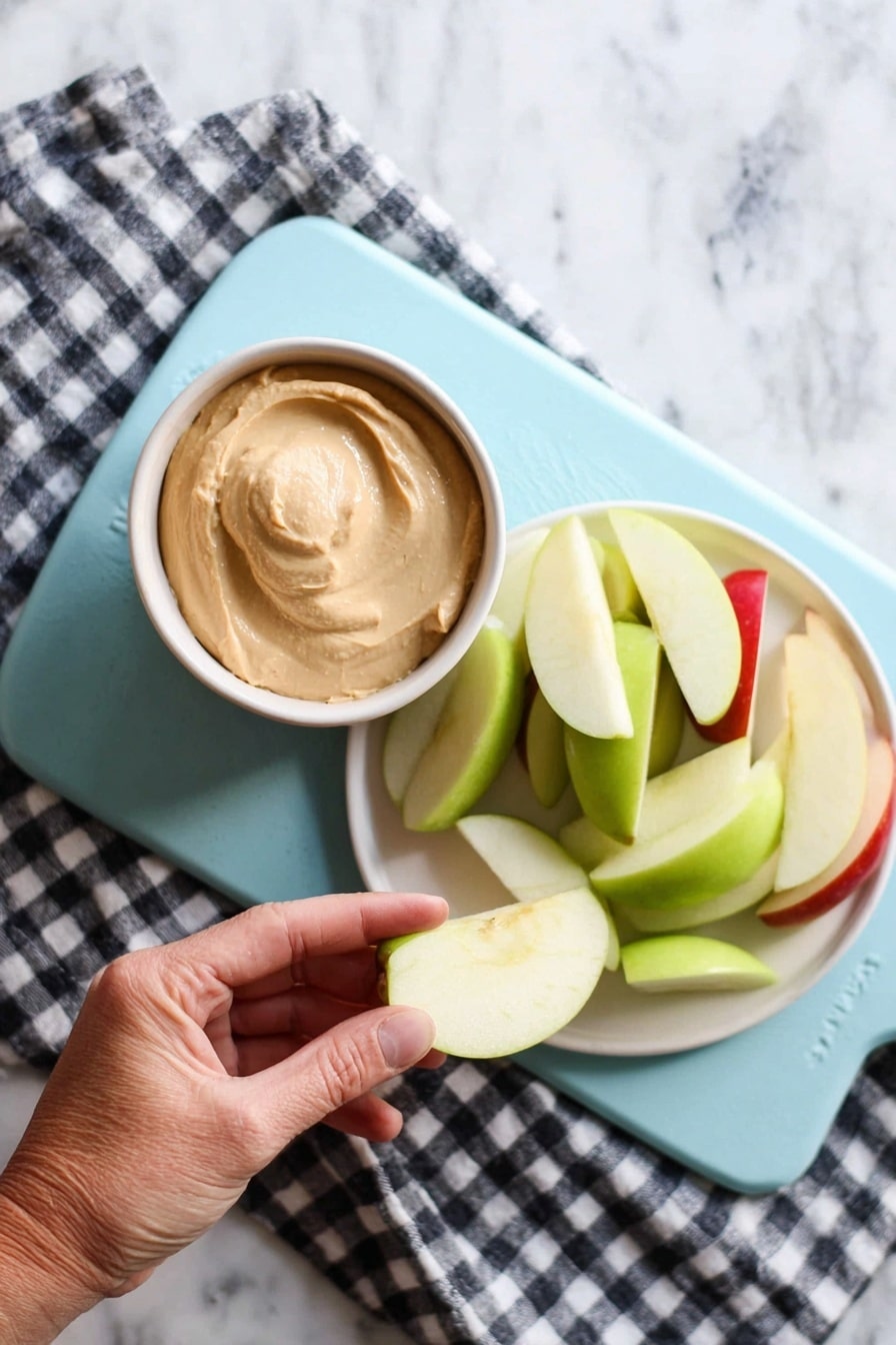 A woman's hand is holding a pale green apple slice dipped in a small bowl of light brown creamy dip. Next to it, on a white plate, there is a pile of apple slices in green and red colors. Both the bowl and plate are placed on a light blue cutting board, which rests on a black and white checkered cloth on a white marbled surface. The scene is brightly lit, showing clear textures of the creamy dip and crisp apple slices. photo taken with an iphone --ar 2:3 --v 7 - Caramel Apple Dip, easy caramel apple dip, best caramel apple dip recipe, quick apple dip, creamy caramel apple dip