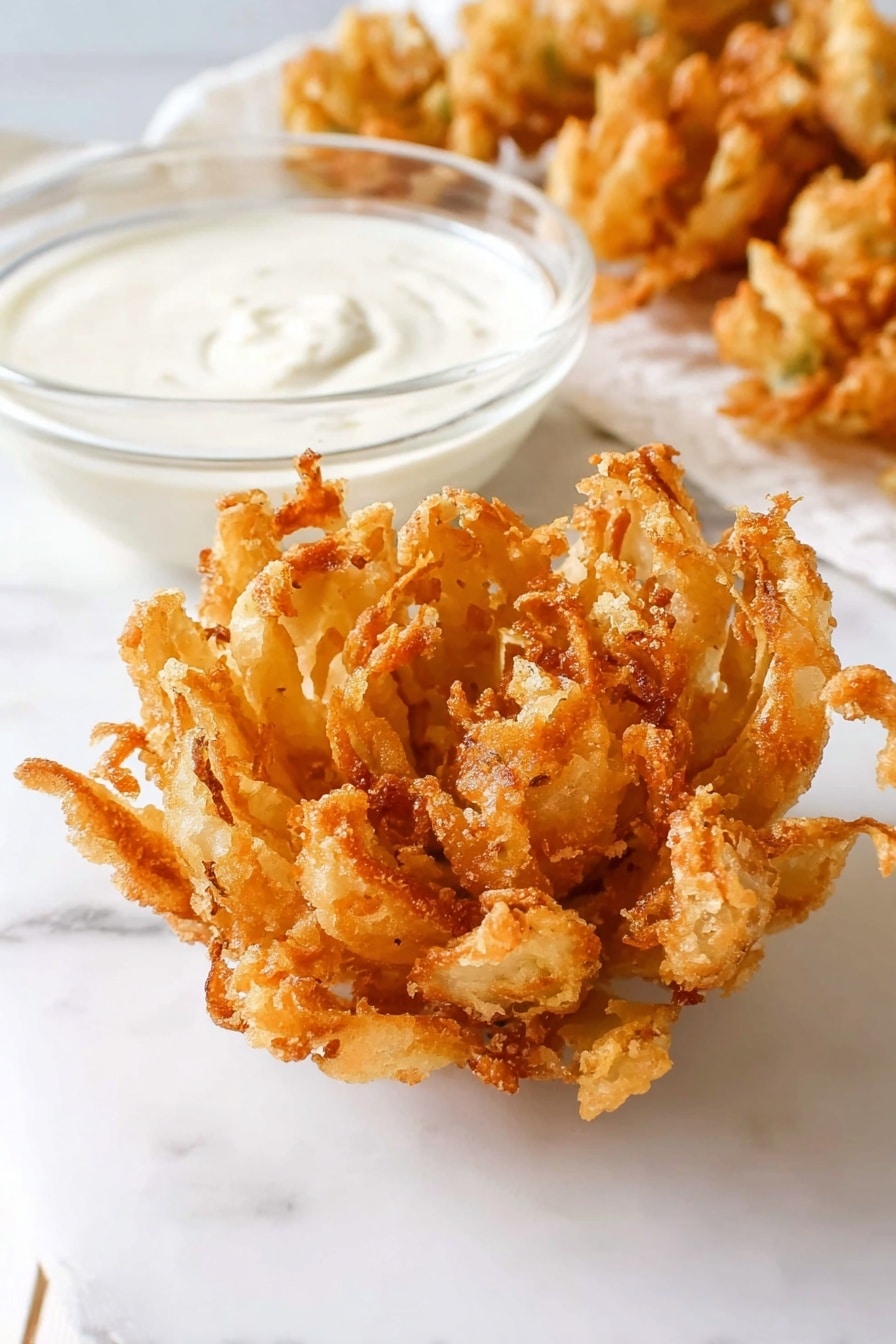 A close-up image of a golden-brown, crispy fried onion blossom placed on a white marbled surface. The onion blossom is fully opened like a flower with many uneven, crunchy petals showing a textured, fried batter coating. Next to it is a small clear glass bowl filled with smooth, creamy white dipping sauce. In the background, some more fried onion blossoms are slightly blurred. The scene is bright and simple with clean white tones and a soft shadow under the food. photo taken with an iphone --ar 2:3 --v 7 - Homemade Blooming Onion, crispy onion appetizer, homemade onion bloom, easy onion appetizer, onion ring recipe