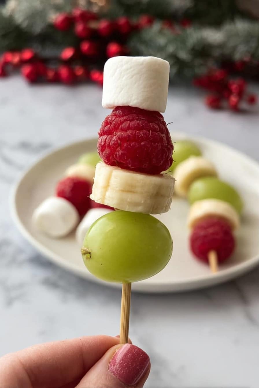 A small skewer held by a woman's hand shows four layers: at the bottom, a smooth, green grape; above it, a round, pale yellow banana slice; next a bright red raspberry with visible bumpy texture; and on top, a small white marshmallow shaped like a cube. In the background, more similar skewers rest on a white plate on a white marbled surface with some red berries. The overall look is fresh, colorful, and festive. photo taken with an iphone --ar 2:3 --v 7 - Grinch Fruit Kabobs, festive fruit skewers, holiday kids snacks, easy healthy party food, colorful holiday treats