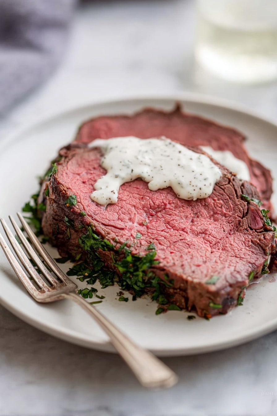 A round thick slice of roast beef with a pink center and brown cooked edges is placed on a white plate. The bottom edge of the beef is covered with chopped green herbs. On top of the beef slice, there is a small amount of white creamy sauce with black specks. A metal fork rests on the left side of the plate. The plate sits on a white marbled surface. Photo taken with an iphone --ar 2:3 --v 7 - Herb Crusted Beef Tenderloin with Horseradish Sauce, beef tenderloin with herb crust, elegant beef main dish, easy roast beef recipe, flavorful horseradish sauce