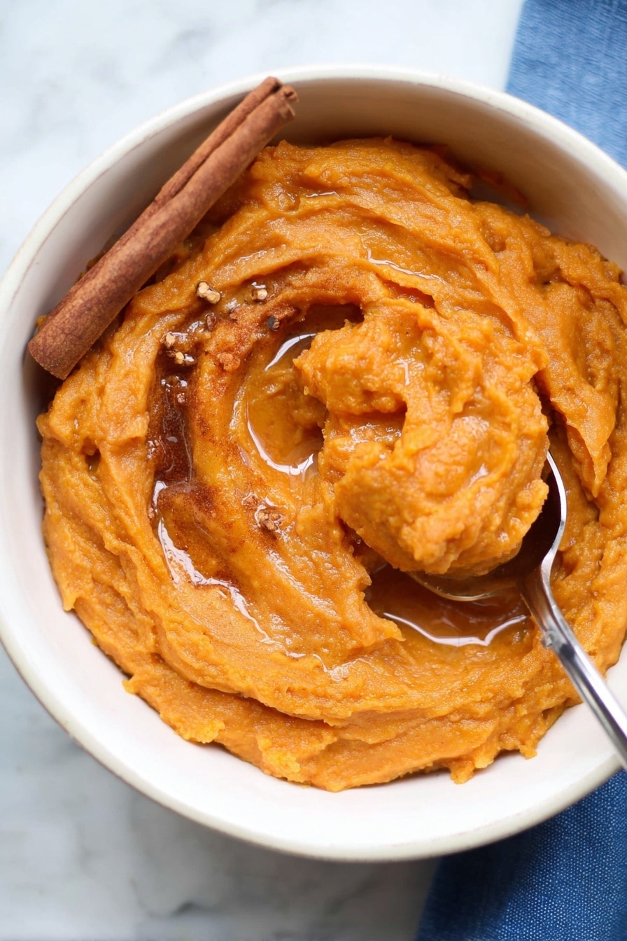 The image shows a white bowl filled with thick, smooth orange sweet potato mash. The mash has swirled texture with a shiny surface, topped with a light drizzle of syrup pooling slightly in the center. A cinnamon stick rests on the left side of the mash, and small bits of brown spices are sprinkled on top. A metal spoon is scooping up part of the mash on the right side, with the spoon handle extending out of the bowl. The bowl sits on a white marbled surface with a bit of a blue cloth visible in the bottom right corner. Photo taken with an iphone --ar 2:3 --v 7 - Brown Butter Sweet Potato Mash, sweet potato mash ideas, cozy side dishes, fall comfort foods, easy sweet potato recipes