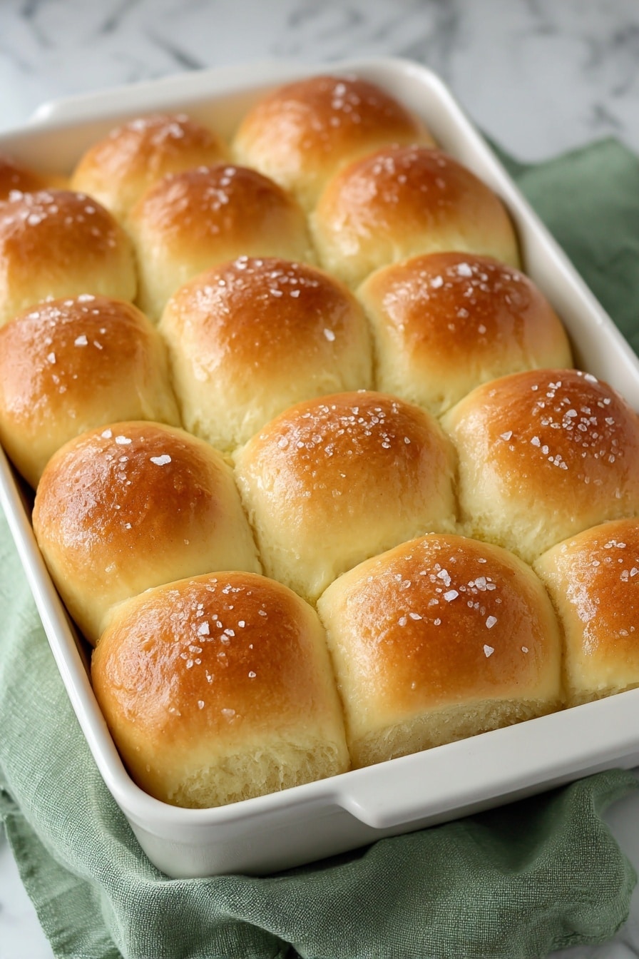 A rectangular white baking pan filled with fifteen golden brown dinner rolls arranged in three rows of five. The rolls have smooth, shiny tops with a soft, slightly puffy texture, and are sprinkled lightly with coarse salt. The baking pan sits on a folded green cloth over a white marbled surface. Photo taken with an iphone --ar 2:3 --v 7 - Sweet Potato Dinner Rolls, sweet potato rolls recipe, fluffy dinner rolls with sweet potatoes, homemade sweet potato rolls, soft bread rolls with sweet potatoes