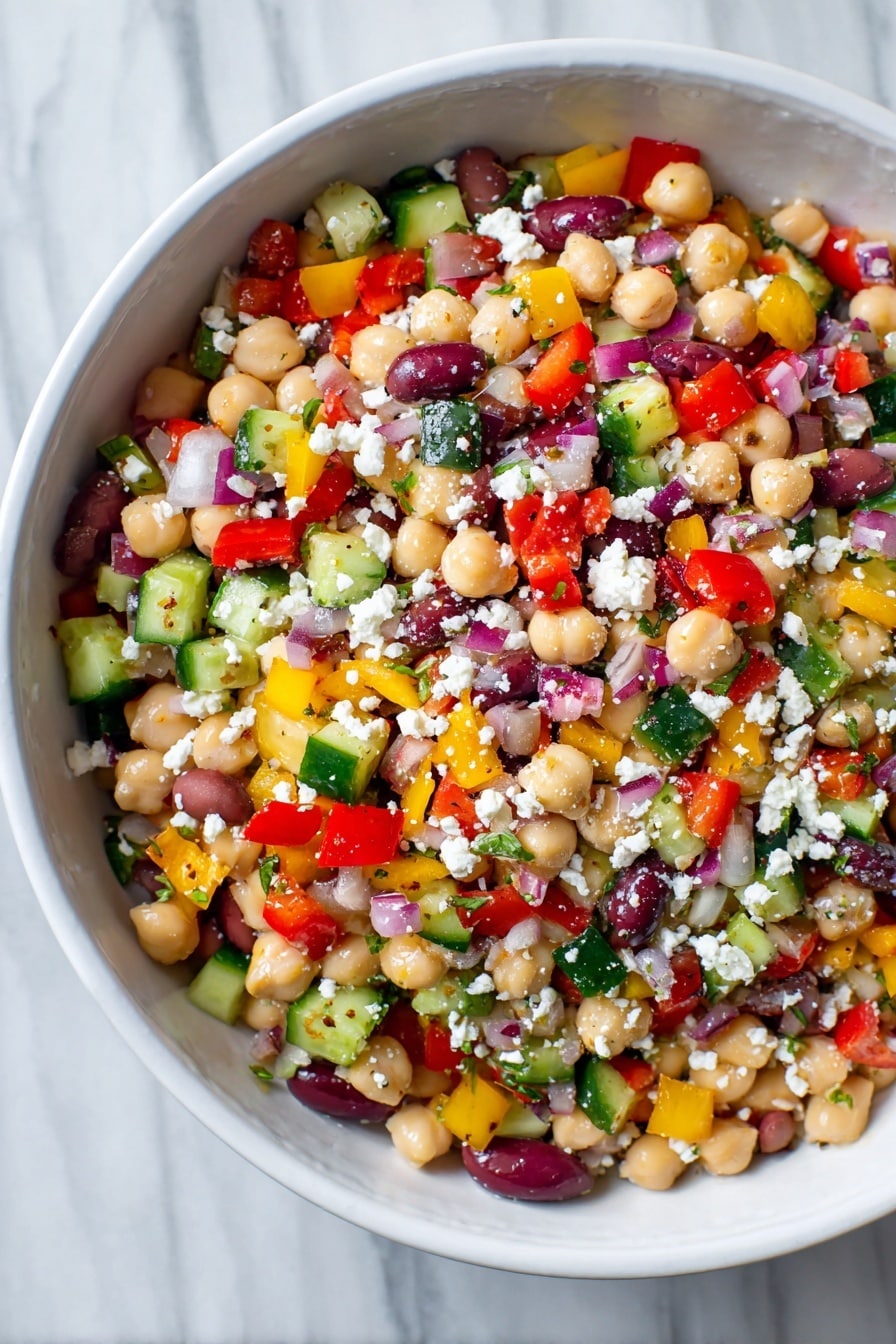 A white bowl filled with a colorful bean salad showing multiple layers of small, chopped vegetables and legumes. The base layer is made of light beige chickpeas and white beans mixed with a scattering of deep purple olive slices. On top, there is a thick mix of small diced bright red bell peppers, yellow bell peppers, green cucumbers, and red onions, creating a vibrant patchwork of colors. White crumbled cheese is sprinkled evenly over the salad, adding a soft, crumbly texture. The surface of the salad looks fresh, moist, and lightly coated in dressing. The bowl rests on a white marbled texture. Photo taken with an iphone --ar 2:3 --v 7 - Hearty Bean Salad with Feta and Olives, healthy bean salad, Mediterranean bean salad, quick vegetarian salad, nutritious side dish