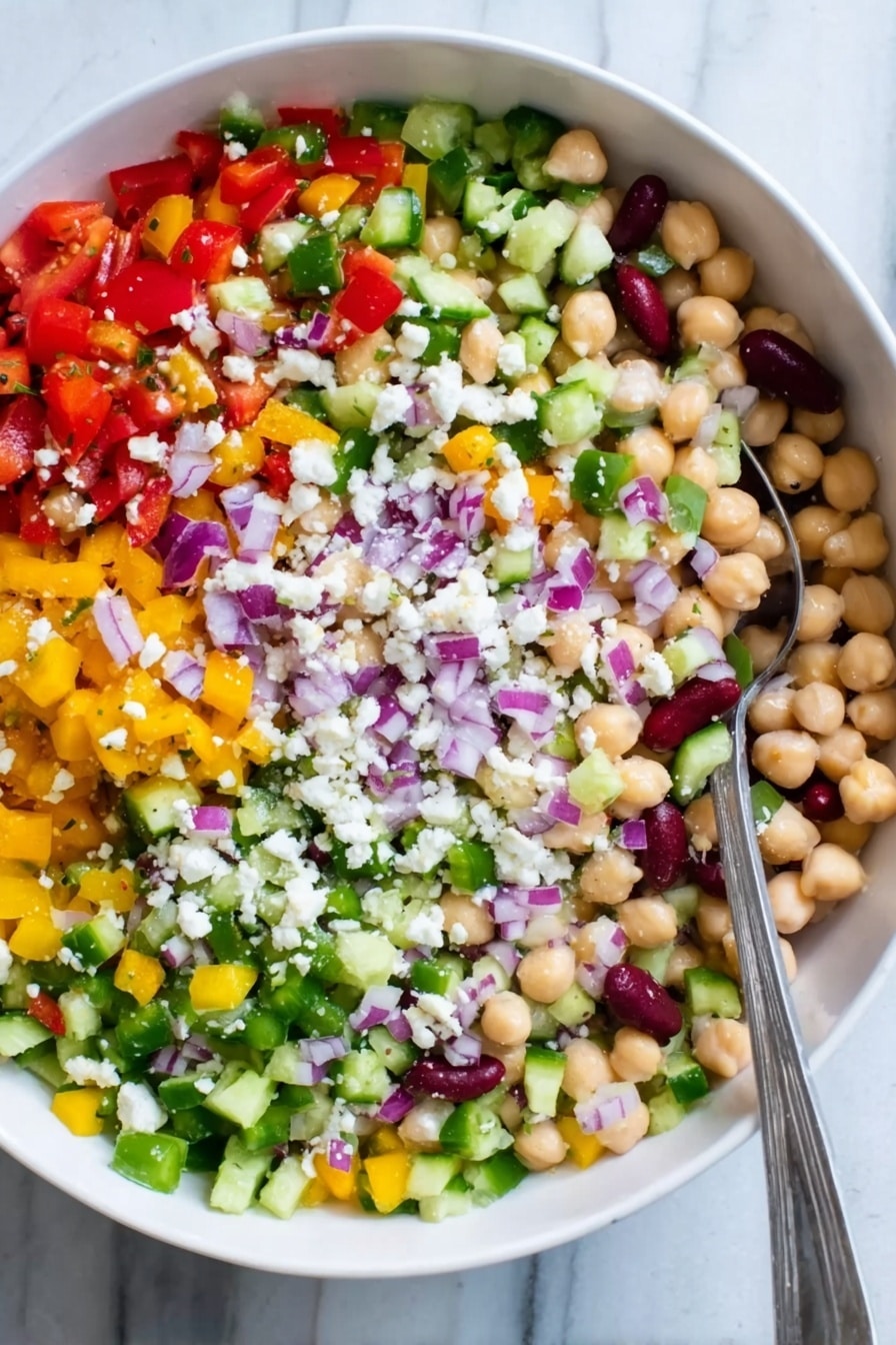 The image shows a white bowl filled with a colorful salad made of many small chopped vegetables and beans. There are five visible layers: at the bottom is a mix of small round chickpeas and white beans with a creamy texture, above that are chopped bright red tomatoes and diced yellow peppers spread evenly, next is a layer of finely chopped green celery and cucumber giving a fresh look, on top of that are small pieces of purple onions scattered across, and sprinkled all over are tiny white crumbles of cheese. A silver spoon is resting on the left side inside the bowl. The bowl sits on a white marbled surface. Photo taken with an iphone --ar 2:3 --v 7 - Hearty Bean Salad with Feta and Olives, healthy bean salad, Mediterranean bean salad, quick vegetarian salad, nutritious side dish