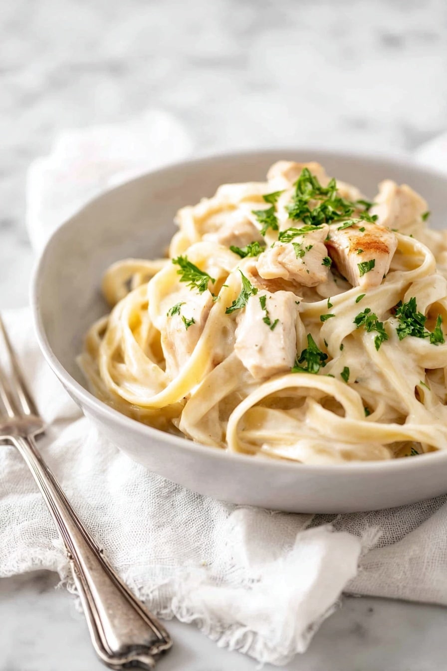 A white bowl holds creamy fettuccine pasta topped with pieces of light golden cooked chicken. The pasta is thick and pale, coated with a smooth white sauce. There are small green bits of parsley sprinkled on top, adding a fresh touch. The bowl sits on a soft white cloth, placed on a white marbled surface. Nearby, a silver fork rests on the cloth. photo taken with an iphone --ar 2:3 --v 7 - One Pot Chicken Alfredo, Chicken Alfredo Pasta, Creamy Chicken Alfredo, Easy Alfredo Pasta, One Pot Pasta Recipes