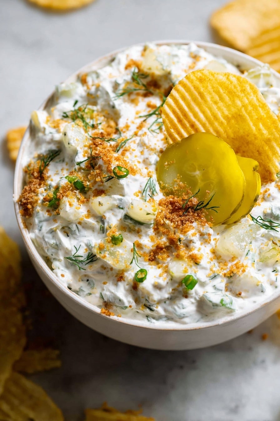 A white bowl filled with a creamy white dip mixed with small green herb pieces and light yellow chunks, topped with bright yellow pickle slices near the center. The dip is sprinkled with light brown crumbs and small green onion pieces, with a few small green herb sprigs scattered on top. A ridged golden potato chip is dipped into the creamy mixture near the edge of the bowl. The bowl sits on a white marbled surface with some crumbs and chips around it. photo taken with an iphone --ar 2:3 --v 7 - Ranch Fried Pickle Dip, fried pickle dip without frying, easy ranch pickle dip, tangy pickle dip recipe, game day dip with pickles