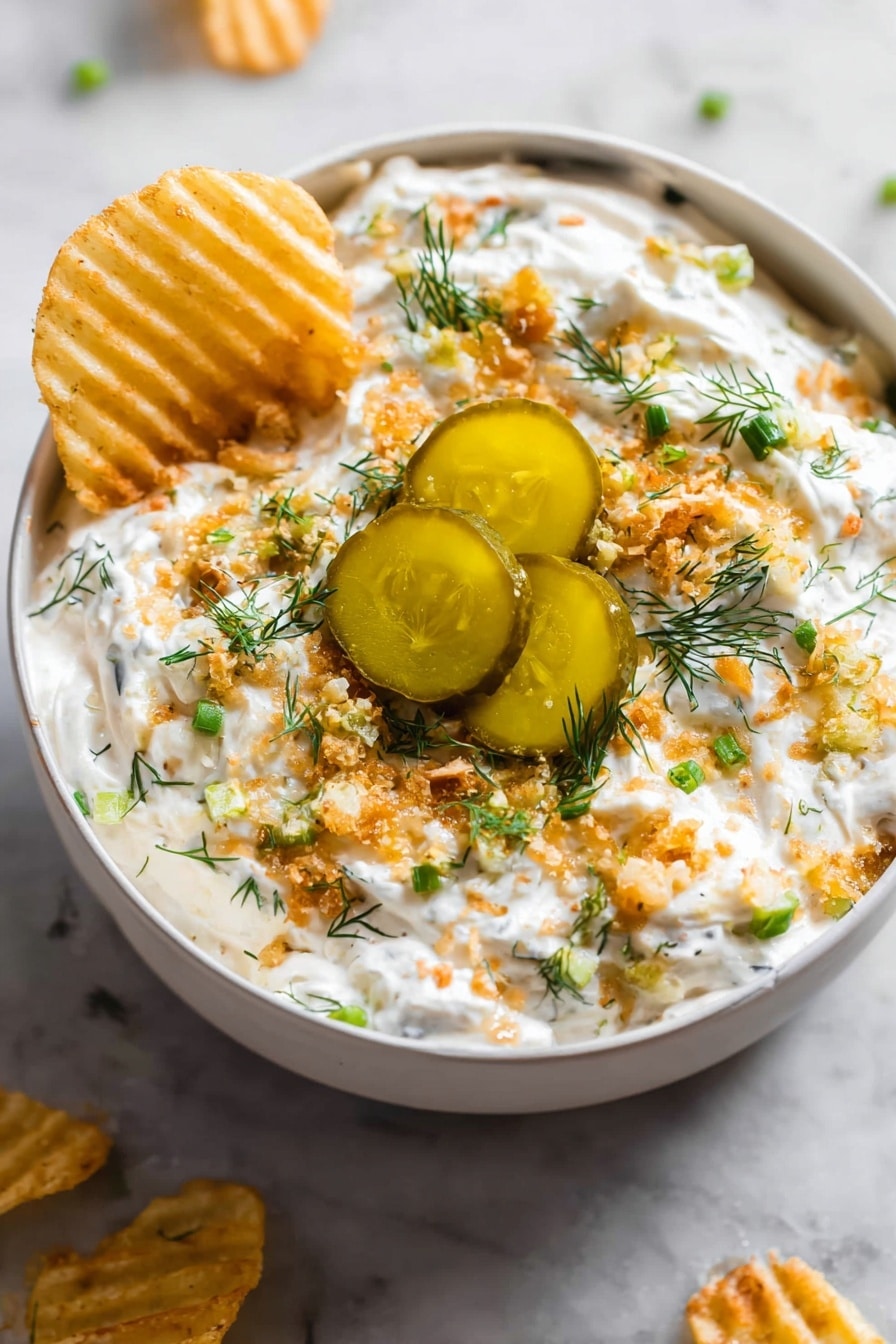 The image shows a grey bowl filled with a creamy white dip that is topped with several layers: small green chopped herbs, light brown crunchy bits, and three thick slices of pickles with a shiny, oily surface in the center. The dip looks thick and textured, with the crunchy bits scattered evenly on top. A woman’s hand is dipping a ridged, yellow chip into the creamy white mixture on one side. The bowl sits on a white marbled surface with some chips placed around it. photo taken with an iphone --ar 2:3 --v 7 - Ranch Fried Pickle Dip, fried pickle dip without frying, easy ranch pickle dip, tangy pickle dip recipe, game day dip with pickles