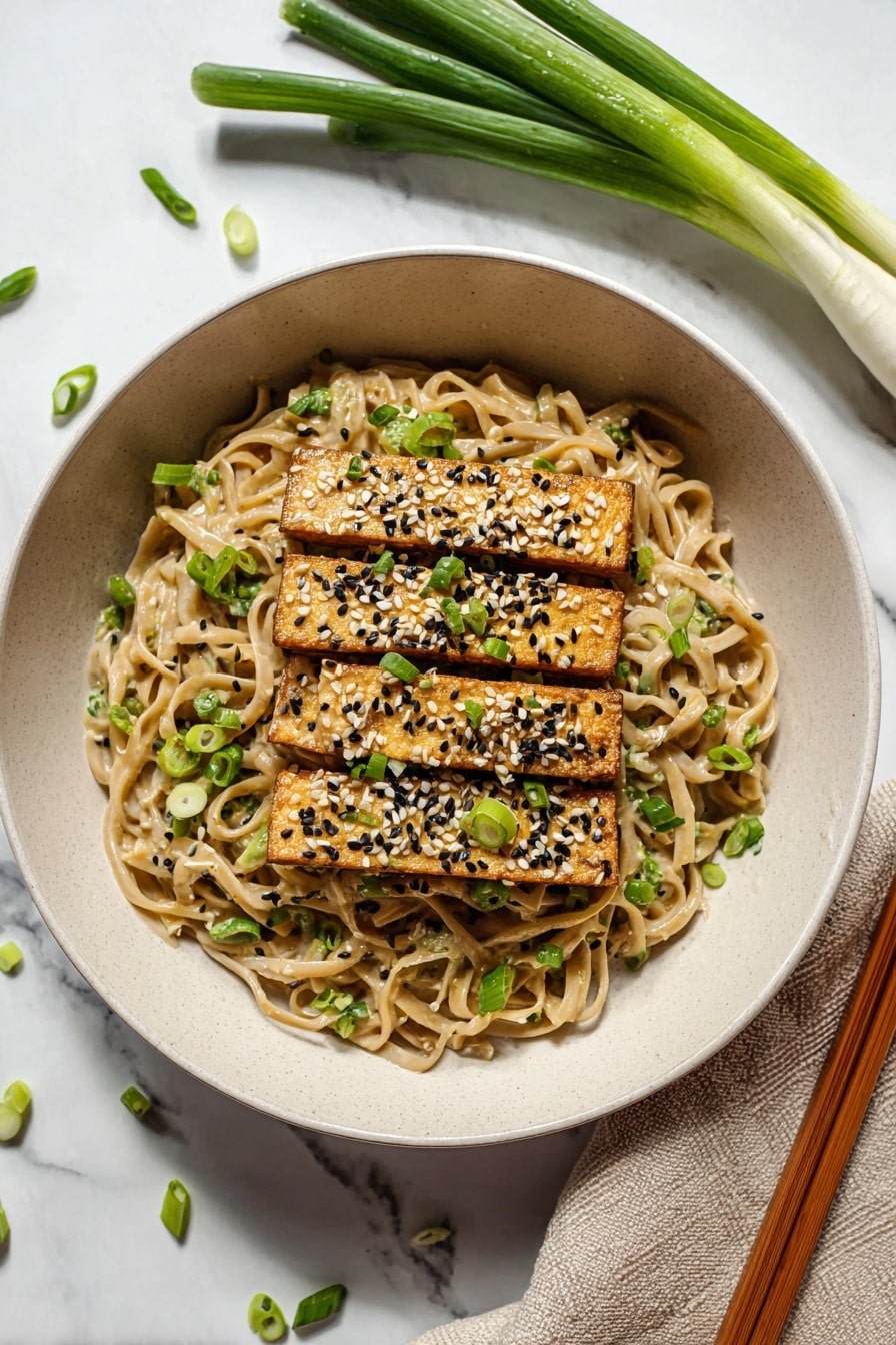 The image shows a close-up of a bowl with three layers. The bottom layer is thick, light tan noodles with a creamy sauce, speckled with tiny black and white seeds. On top of the noodles, there are golden-brown rectangular tofu pieces sprinkled with white and black sesame seeds and chopped green onions. At the top, a pair of wooden chopsticks held by a woman's hand lifts some noodles, with a few green onion slices stuck to the chopsticks. The bowl is white, and the background surface has a white marbled texture. photo taken with an iphone --ar 2:3 --v 7 - Peanut Udon Noodles, Thai peanut udon, quick udon noodle recipe, vegan peanut noodles, easy Asian noodle dish