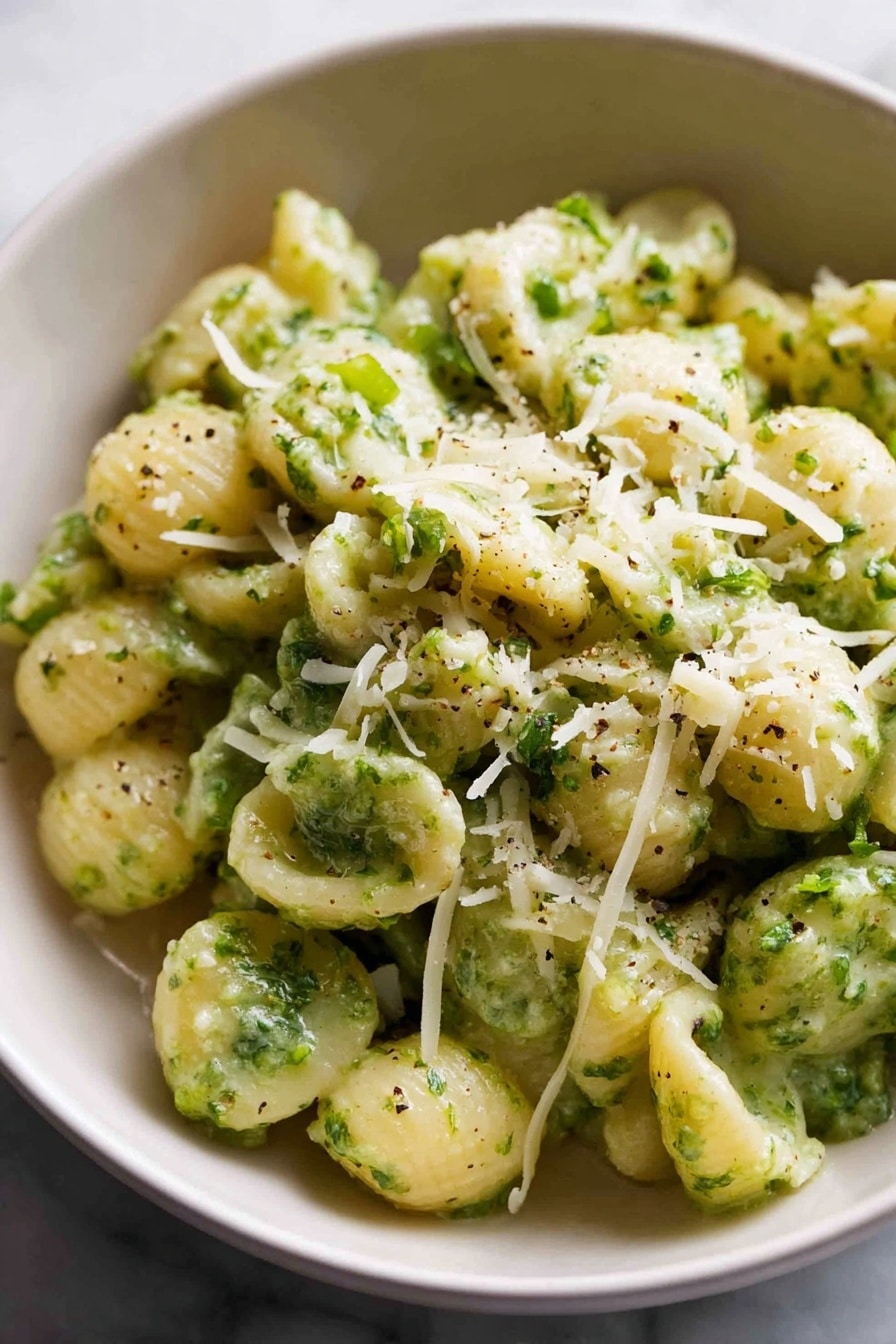 A large bowl filled with small spiral pasta mixed with bright green broccoli florets. The pasta is light yellow and looks soft, while the broccoli pieces are spread evenly throughout the dish. On top, there is a sprinkling of grated parmesan cheese, adding a slight white textured layer. A silver spoon is placed inside the bowl on the left side, ready to serve. In the background, there are two stacked white bowls, a silver fork and spoon set on a white cloth, and a block of parmesan cheese next to a grater on a white marbled surface. The overall setting feels cozy and inviting. photo taken with an iphone --ar 2:3 --v 7 - Quick Broccoli Pasta with Lemon and Cheese, easy broccoli pasta recipe, quick lemon broccoli pasta, healthy cheesy pasta, 15-minute pasta dinner