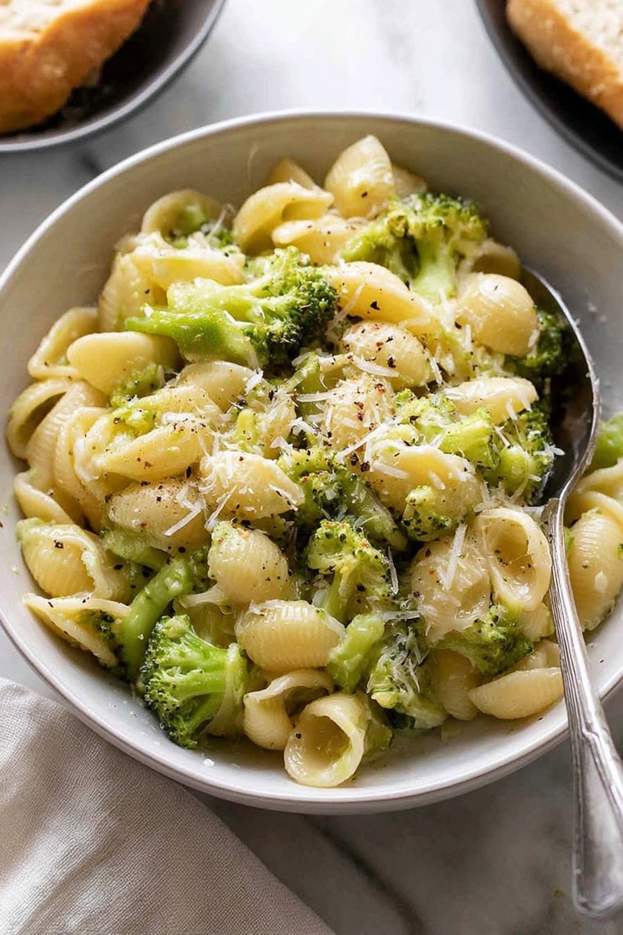 The image shows a white bowl filled with small shell pasta mixed with bright green broccoli pieces. The pasta looks soft and is coated with a light creamy sauce that makes the dish look smooth and shiny. There is a sprinkle of finely grated cheese on top and tiny specks of black pepper scattered over the dish. A silver spoon is placed partially inside the bowl on the right side. The bowl is set on a white marbled surface with a light cloth nearby and a blurred part of another dish with bread in the background. Photo taken with an iphone --ar 2:3 --v 7 - Quick Broccoli Pasta with Lemon and Cheese, easy broccoli pasta recipe, quick lemon broccoli pasta, healthy cheesy pasta, 15-minute pasta dinner