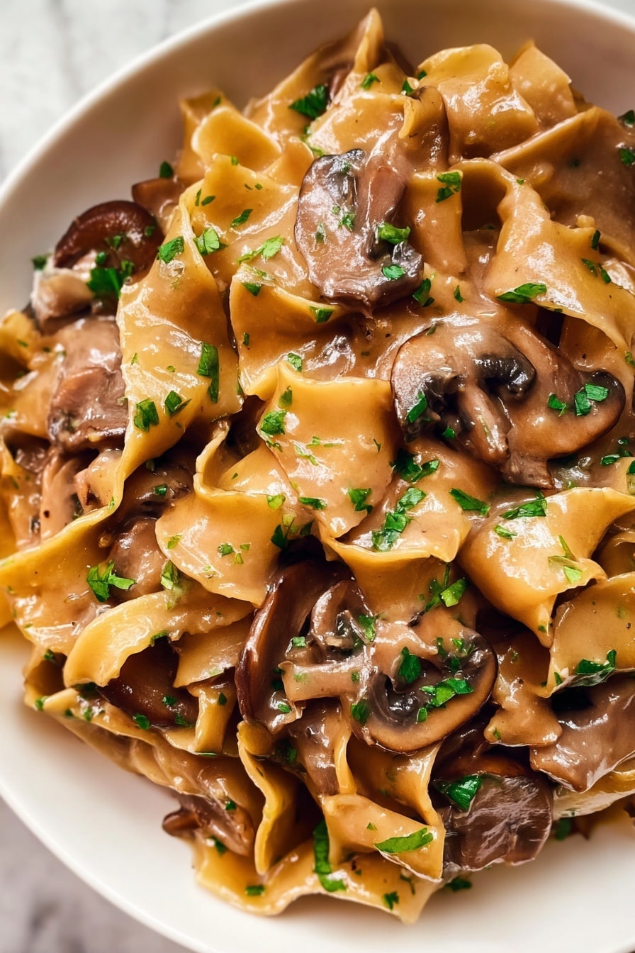 The image shows a close-up of a creamy pasta dish with wide, flat egg noodles mixed with thick slices of brown mushrooms. The noodles are coated in a smooth, light brown sauce that looks rich and glossy. Small pieces of chopped green herbs are sprinkled over the top, adding a fresh touch to the dish. The textures of the soft noodles and tender mushrooms blend together, showing a warm and comforting meal. The dish is served in a white bowl placed on a white marbled surface. photo taken with an iphone --ar 2:3 --v 7 - Vegetarian Mushroom Stroganoff, easy vegetarian stroganoff, creamy mushroom pasta, plant-based stroganoff recipe, quick mushroom dinner
