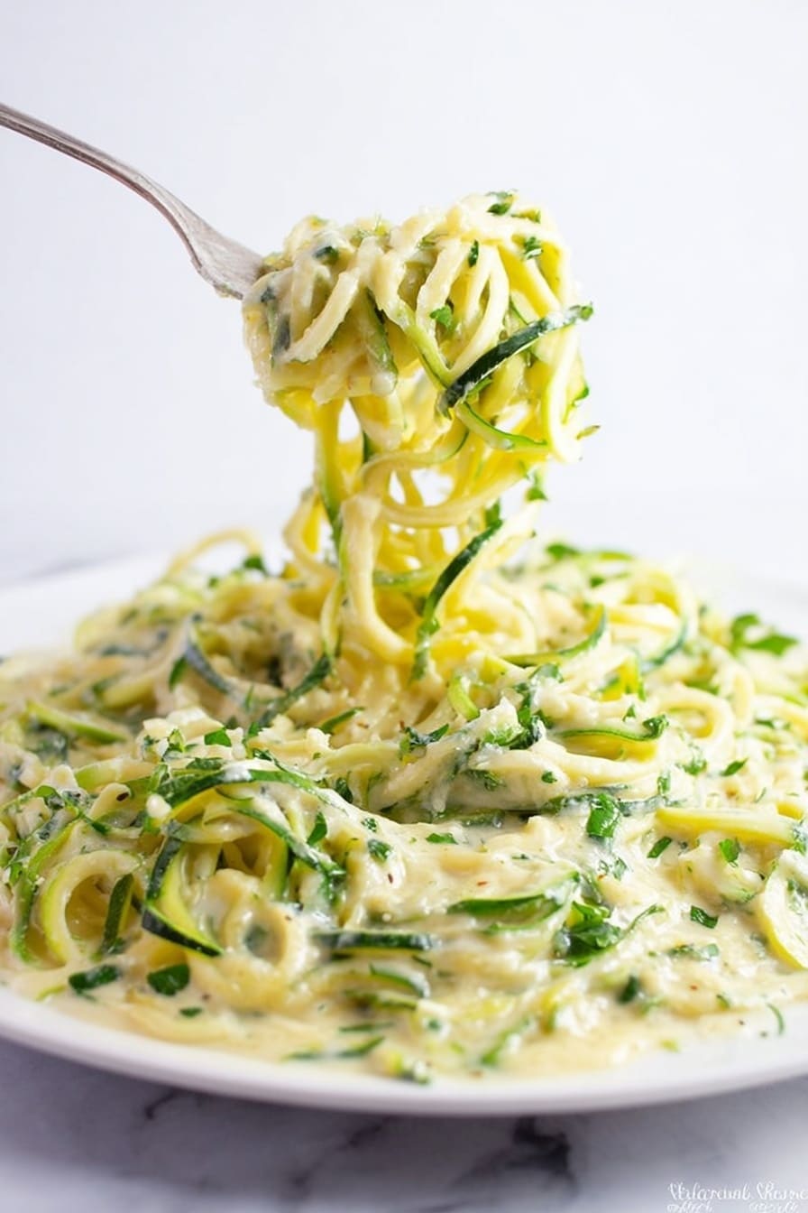 A close-up view of a large white plate filled with spiralized zucchini noodles mixed with a creamy light yellow sauce and sprinkled with small pieces of chopped green herbs, mostly parsley or cilantro. A fork lifts a twirl of the noodles above the plate, showing the long, thin, curly strands coated evenly in the sauce. The background is plain white marbled texture, making the colors of the zucchini and sauce stand out clearly. photo taken with an iphone --ar 2:3 --v 7 - Zucchini Alfredo, healthy zucchini Alfredo, low carb Alfredo with zucchini, creamy zucchini pasta, quick Zucchini Alfredo dinner
