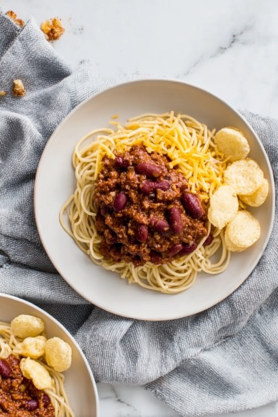 The image shows a white plate with three main layers. The bottom layer is a pile of light brown spaghetti noodles arranged in a loose small mound. On top of the noodles, there is a thick layer of dark reddish-brown chili with visible kidney beans and a chunky texture. To one side of the noodles and chili, there is a small group of round, pale beige crunchy chips neatly placed. The noodles and chips sit on a white marbled surface with a soft grey cloth draped around the plate. In the corner of the photo, part of another similar plate with the same food is visible. Photo taken with an iphone --ar 2:3 --v 7 - Cincinnati Chili Spaghetti, Cincinnati chili, easy chili recipes, hearty spaghetti dish, Ohio comfort food