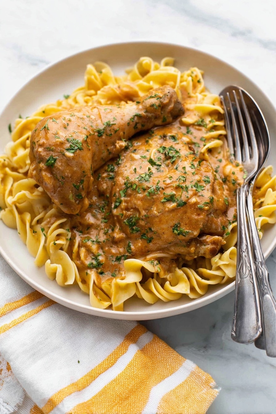 A white round plate holds two pieces of cooked chicken, one drumstick and one thigh, both covered in a thick light brown sauce with green herbs sprinkled on top. Underneath, there is a bed of light yellow egg noodles twisted softly, also coated lightly with the same sauce. A silver fork and spoon rest on the right edge of the plate. The plate sits on a white marbled surface with a white and yellow striped cloth napkin partially visible at the bottom left corner. Photo taken with an iphone --ar 2:3 --v 7 - Authentic Chicken Paprikash, Hungarian Chicken Paprikash, creamy paprika chicken dish, traditional Hungarian chicken recipe, easy Chicken Paprikash