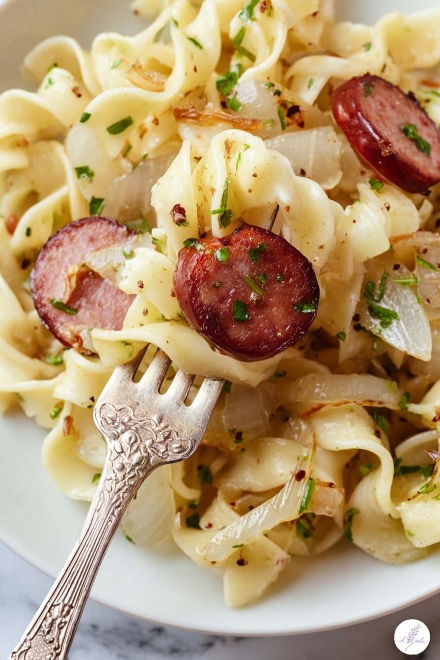 A close-up view of a white plate filled with twisted egg noodles that are light yellow with a soft texture, mixed with small pieces of cooked onions that are translucent white with a slight brown edge, and slices of browned sausage that are dark red with a grilled surface and visible herbs. The noodles and ingredients are sprinkled with chopped green herbs and black pepper. A vintage silver fork with floral engravings is lifting some noodles and one slice of sausage. The background is a white marbled surface. Photo taken with an iphone --ar 2:3 --v 7 - Easy Kielbasa and Cabbage Haluski, kielbasa and cabbage skillet, hearty cabbage and sausage dish, quick kielbasa cabbage dinner, simple haluski recipe