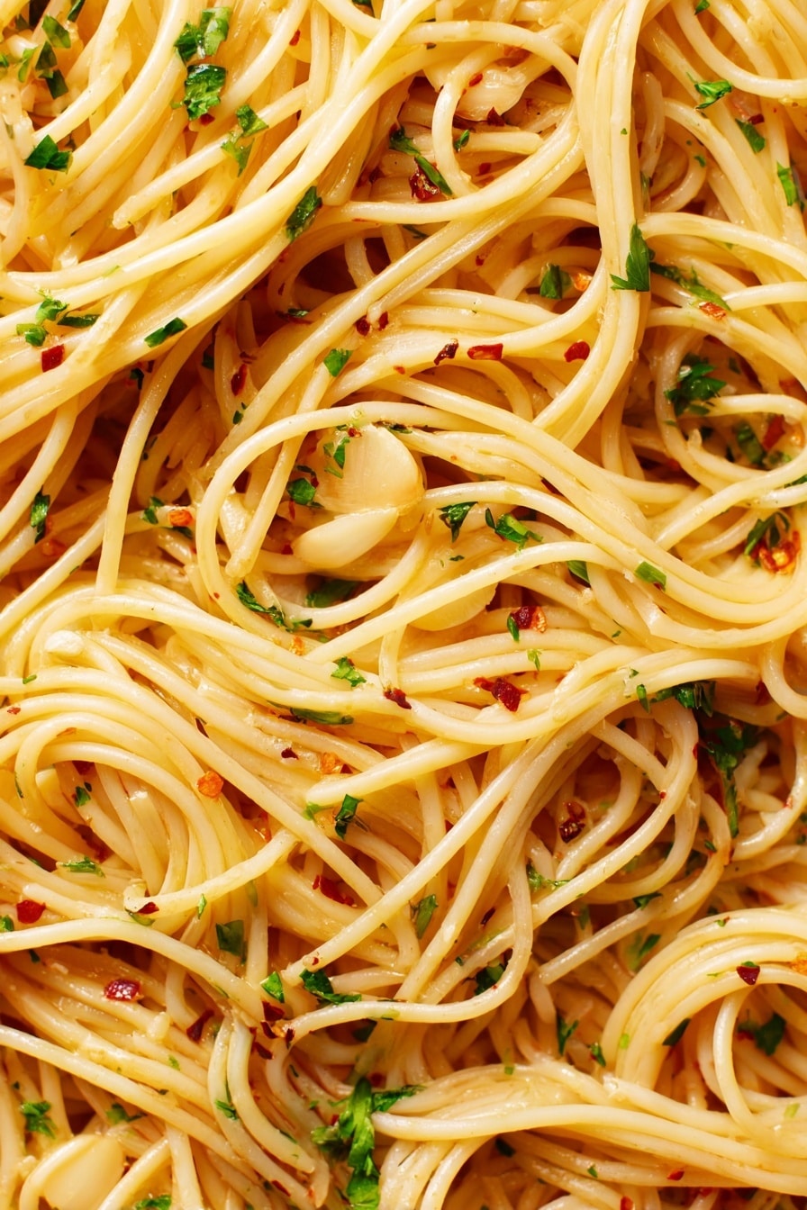 The image shows a close-up view of spaghetti pasta with thin, long noodles that are light golden in color. The noodles are mixed evenly with thin slices of light tan garlic, small bits of green parsley, and tiny red chili flakes scattered throughout. The spaghetti appears glossy with a slightly oily texture, making the noodles look smooth and shiny. The background is a white marbled texture. photo taken with an iphone --ar 2:3 --v 7 - Spaghetti Aglio e Olio, Italian pasta recipes, quick dinner ideas, garlic pasta, easy Italian main dish