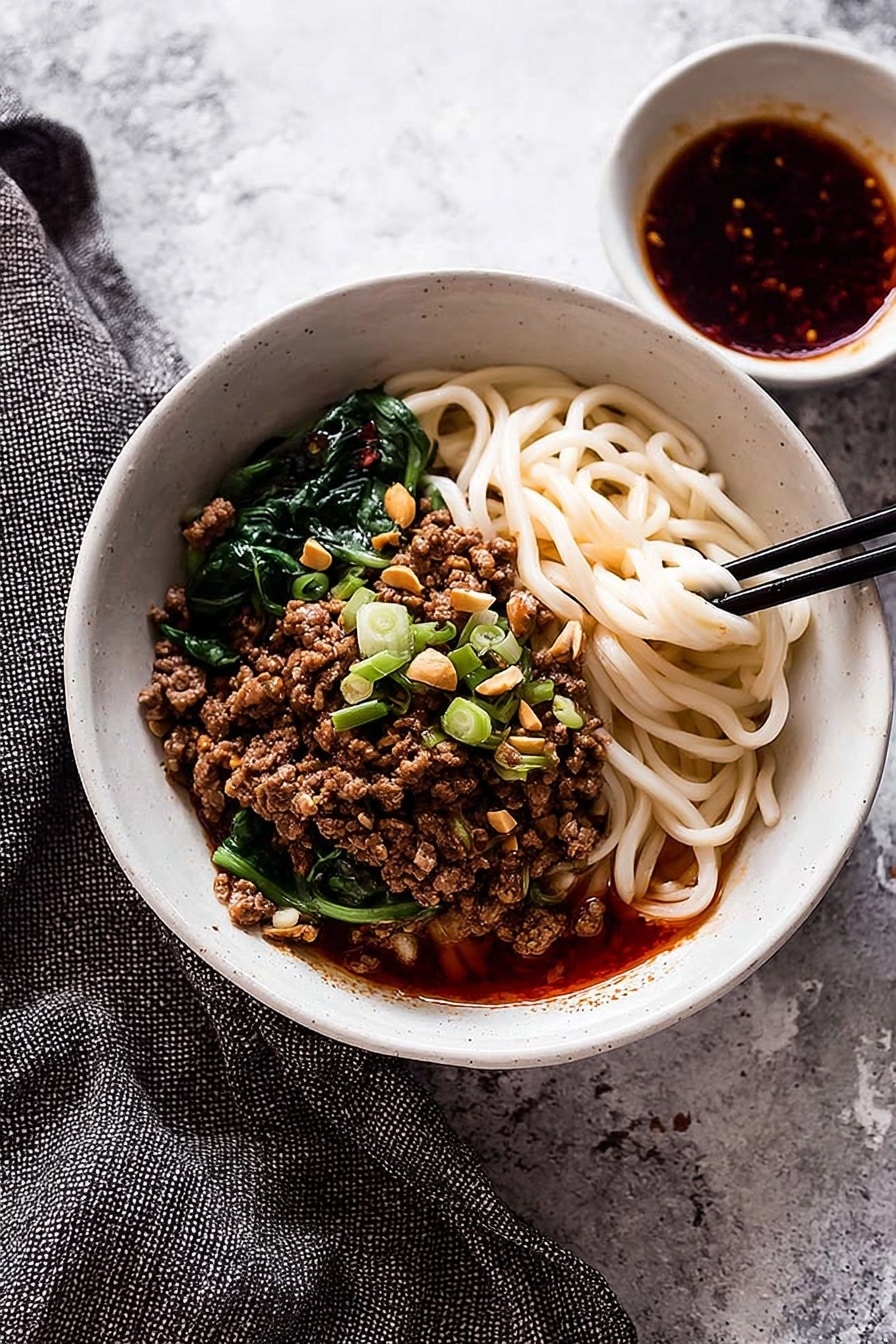 A white bowl filled with three main layers: at the bottom, thick, white noodles partially covered with a red oily sauce; in the middle, a generous layer of browned minced meat topped with chopped green onions and crushed peanuts; and on the top, fresh green leafy vegetables arranged on one side. A pair of wooden chopsticks held by a woman's hand is lifting some noodles, showing their texture and sauce coating. The bowl is placed on a white marbled surface with scattered green onion pieces and crushed peanuts nearby. photo taken with an iphone --ar 2:3 --v 7 - Spicy Dan Dan Noodles with Pork, Sichuan Dan Dan Noodles, Homemade Dan Dan Noodles, Pork and Chili Oil Noodles, Authentic Sichuan Noodles