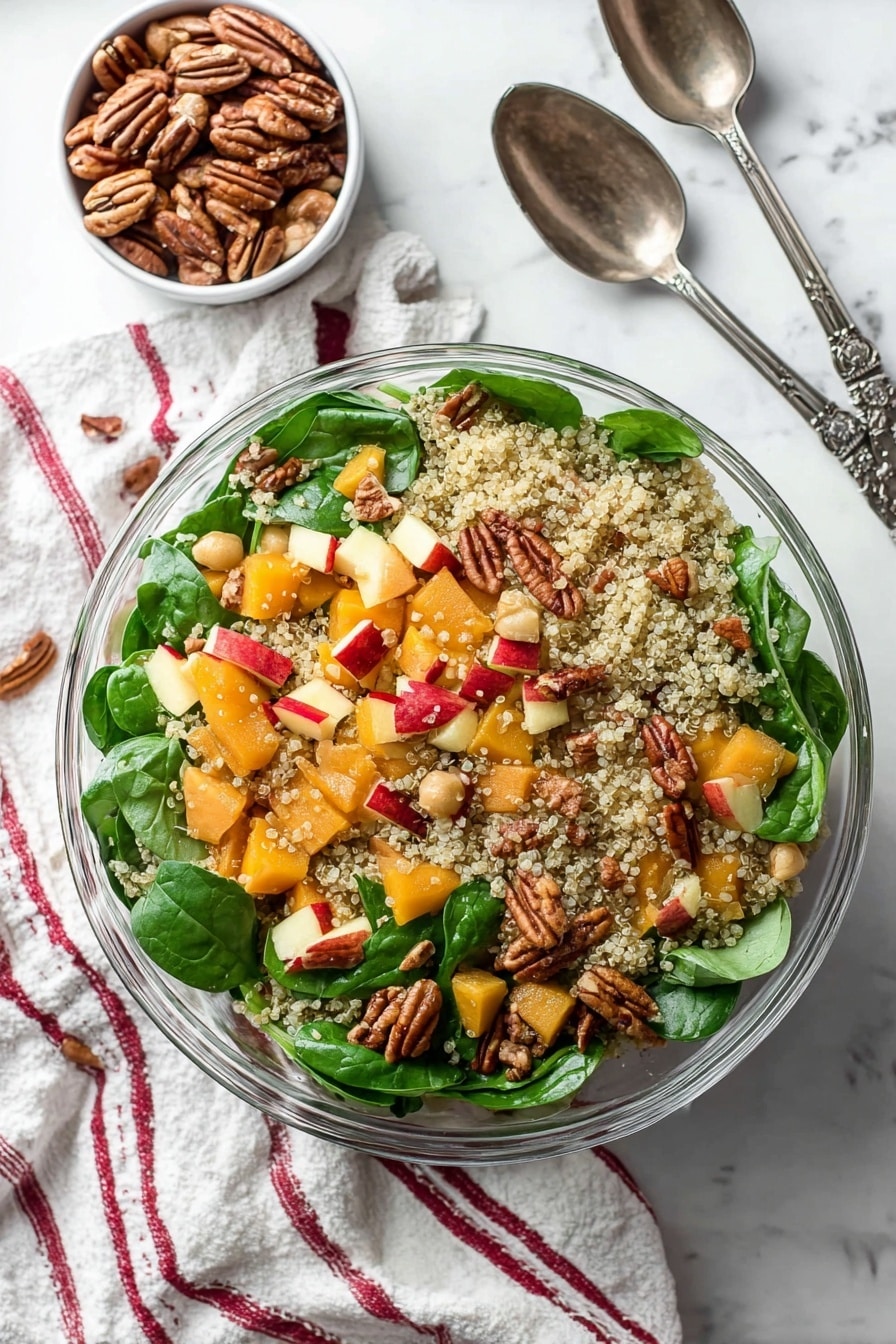 This image shows a clear glass bowl filled with a colorful quinoa salad. The base layer is light, fluffy quinoa with small, round grains scattered evenly throughout. Mixed inside are bright green fresh spinach leaves with a smooth texture, orange chunks of butternut squash, and small pale beans adding softness. Red-skinned apple pieces give a splash of red color, and whole pecans add a textured, brown, nutty layer randomly placed on top. The bowl is on a white marbled surface with a soft striped cloth nearby, and a woman's hand holds a dark metal salad spoon at the edge. photo taken with an iphone --ar 2:3 --v 7 - Roasted Butternut Squash Quinoa Salad, healthy butternut squash salad, autumn quinoa salad, easy roasted squash salad, gluten-free quinoa salad